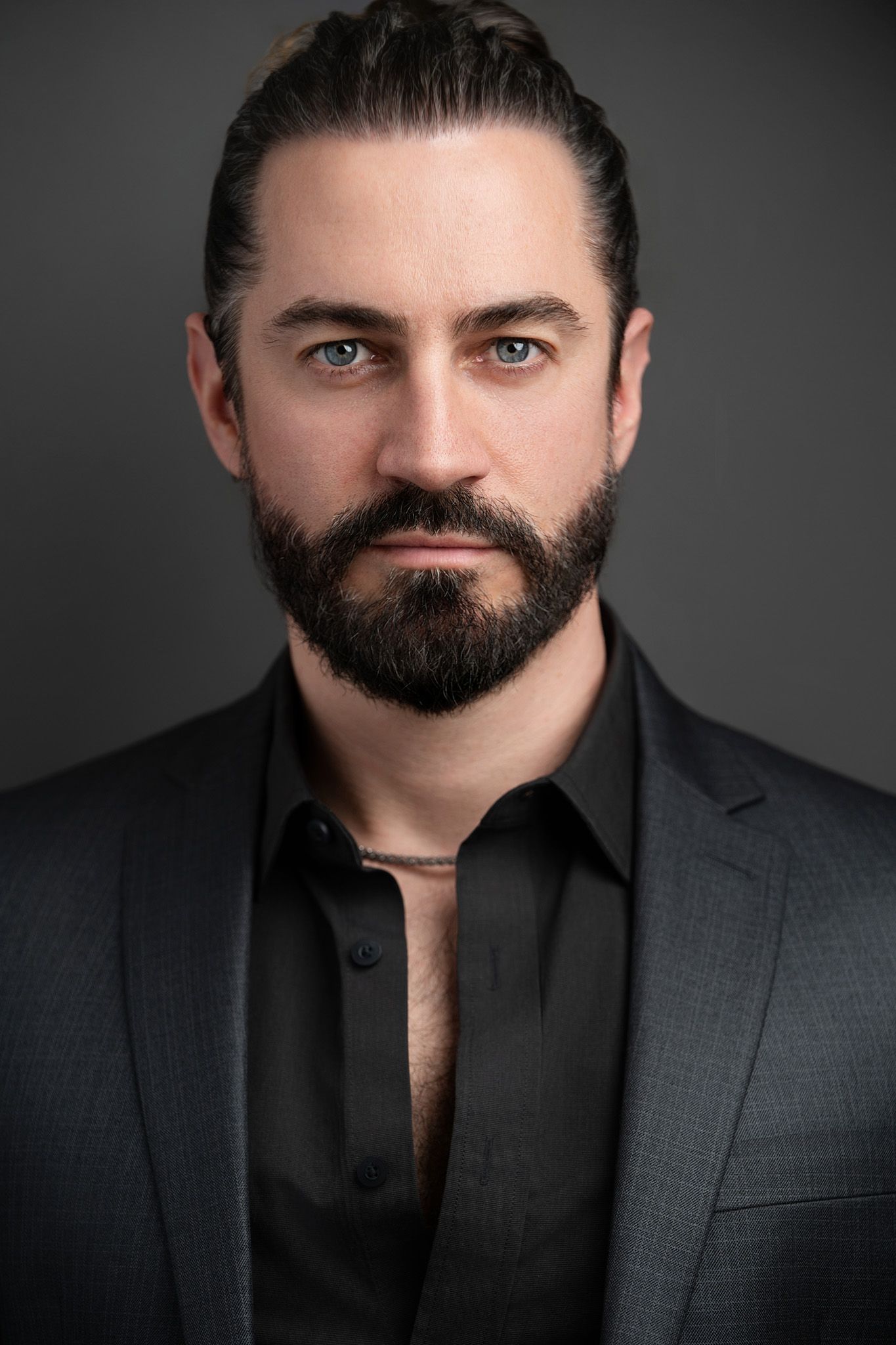 Professional headshot of a bearded man with blue eyes wearing a dark suit and black shirt against gray background.