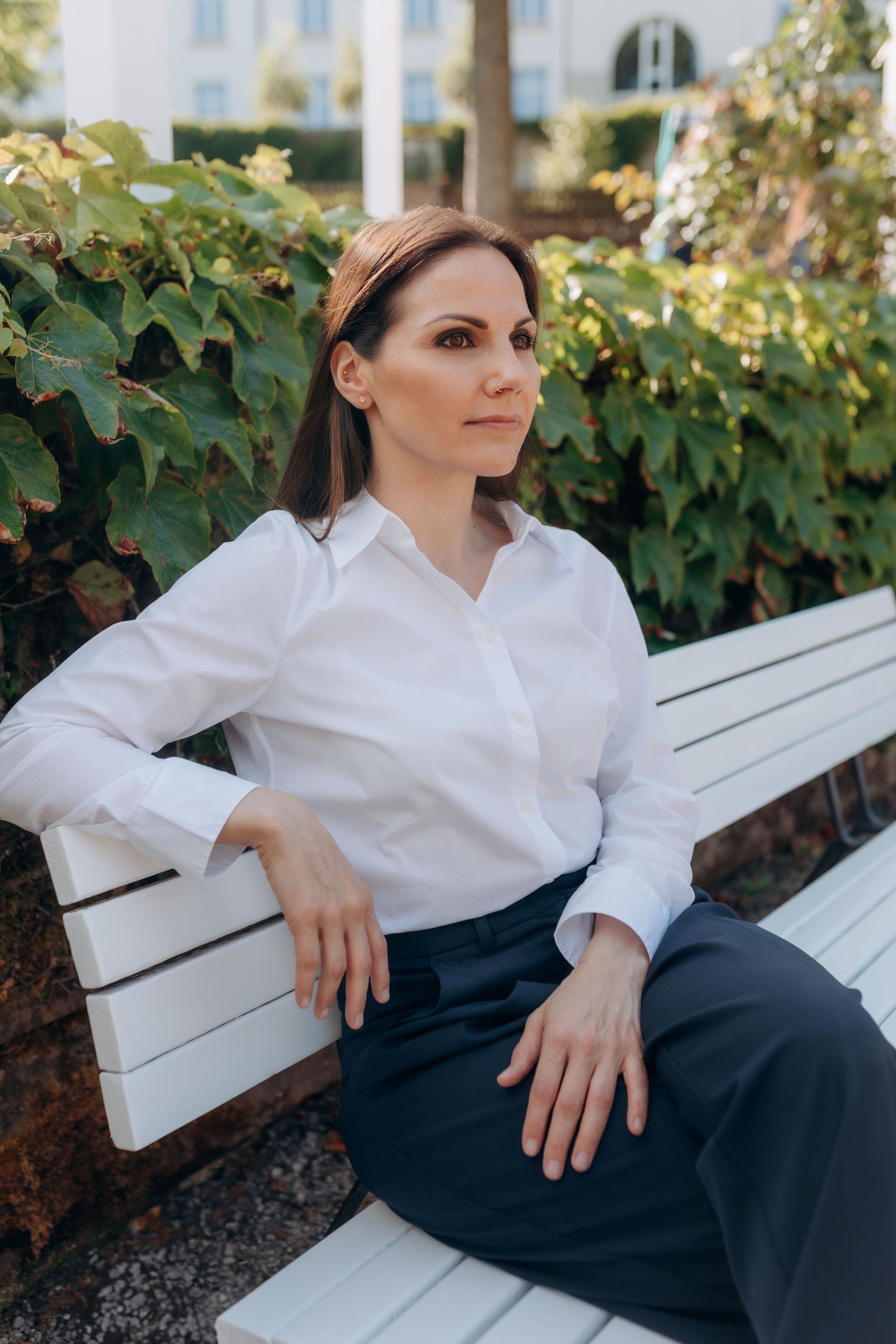 Person wearing crisp white blouse and dark pants rests elegantly on a white bench outdoors with lush greenery in background.