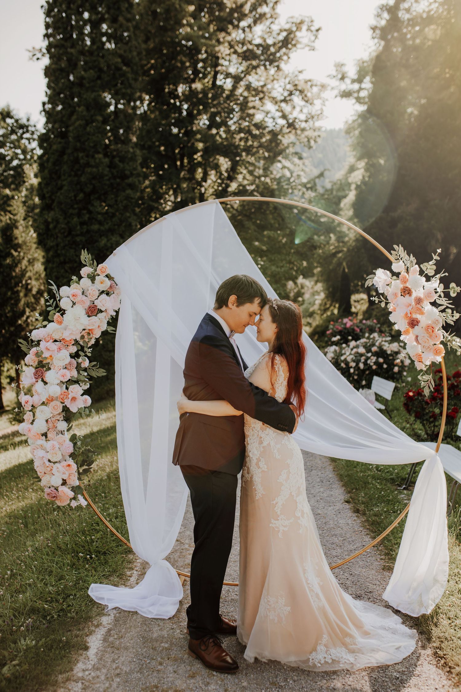 Wedding ceremony setup with a circular arch decorated with pink flowers and white fabric in a romantic outdoor garden setting.