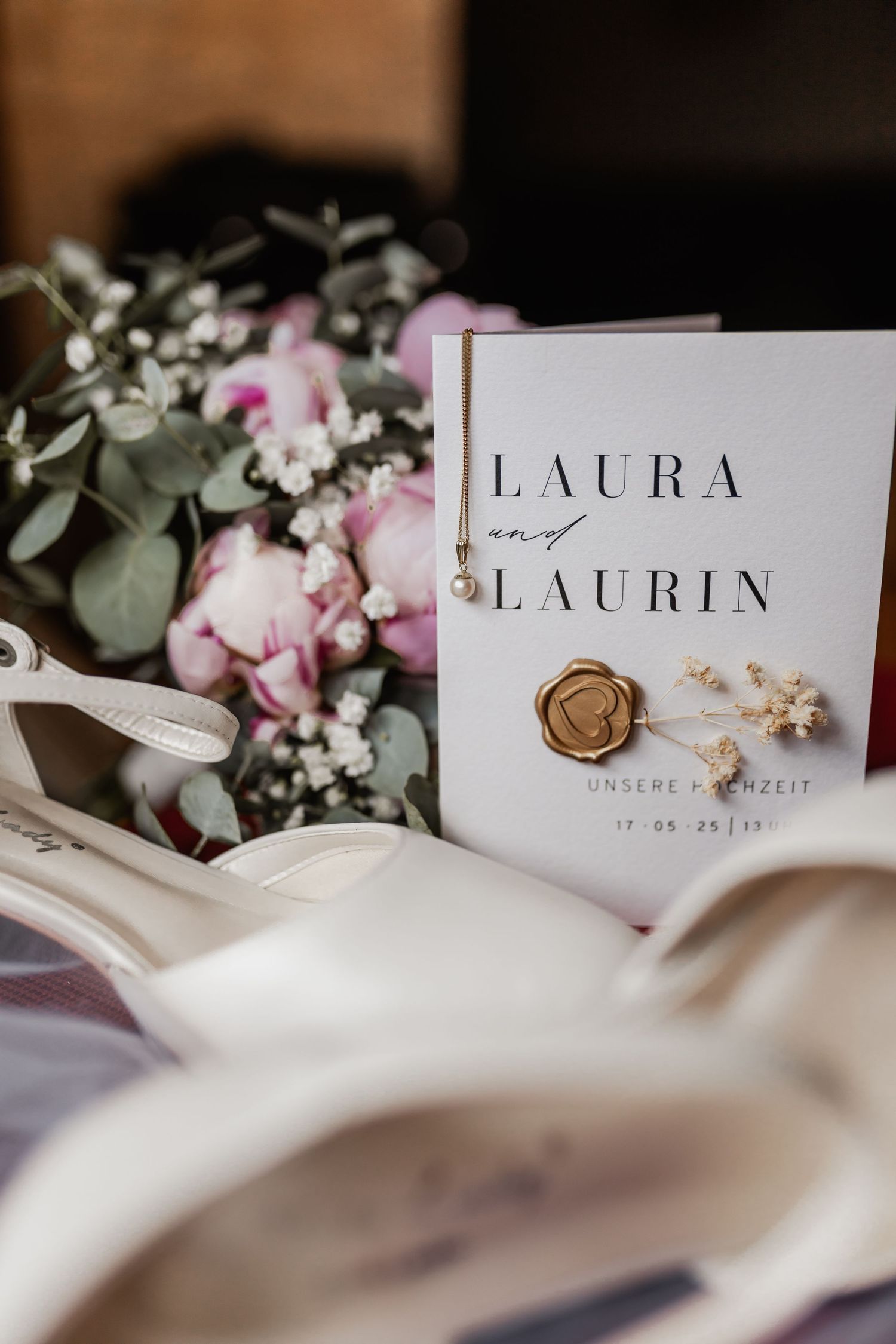 Wedding invitation with names Laura and Laurin displayed alongside pink roses, eucalyptus and a wax seal on white fabric.