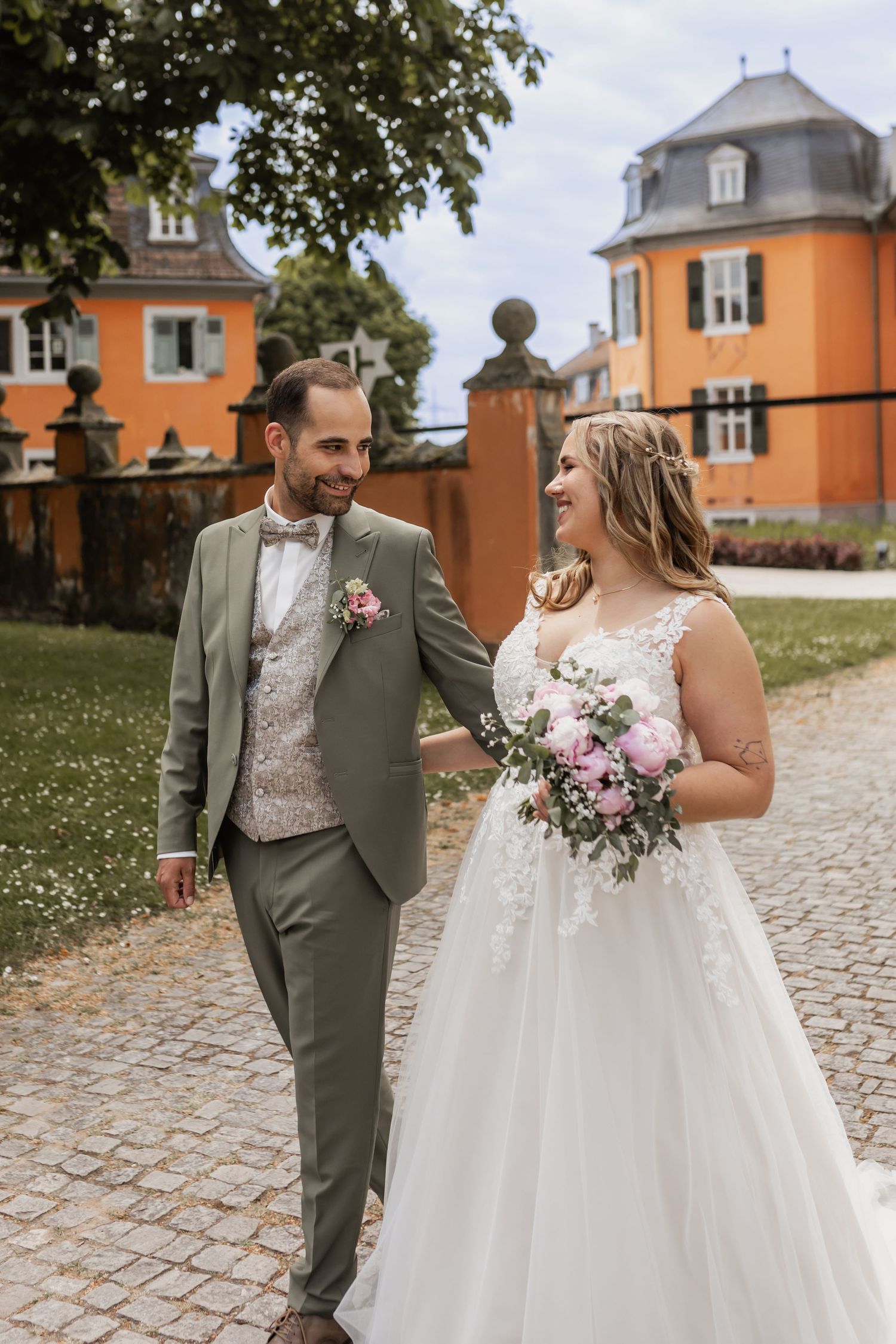 Couple in formal wedding attire walk hand in hand along a cobblestone path beside orange buildings on a sunny day.