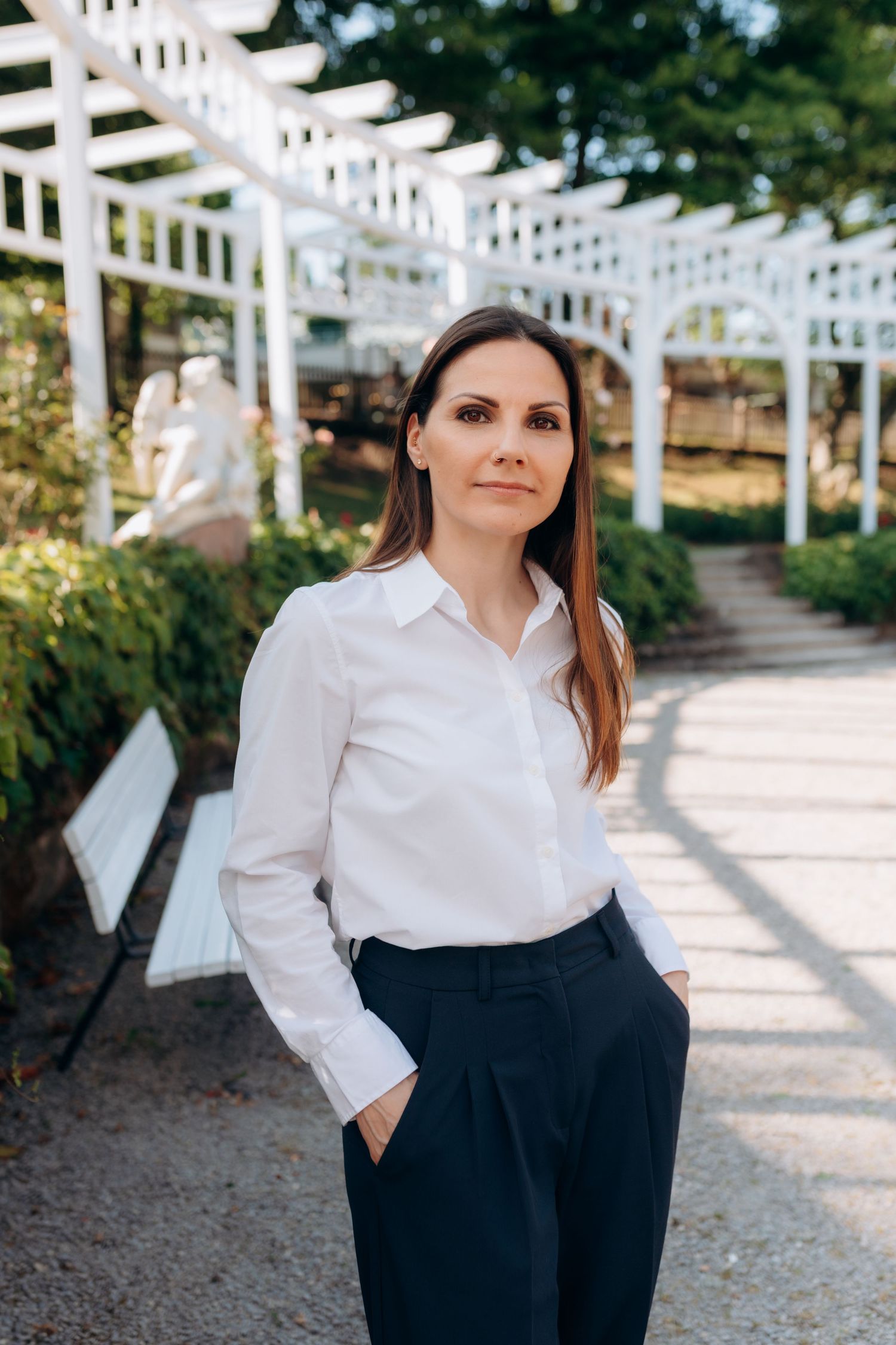 Professional portrait in outdoor garden setting with white lattice pergola, wearing classic white shirt and navy pants.