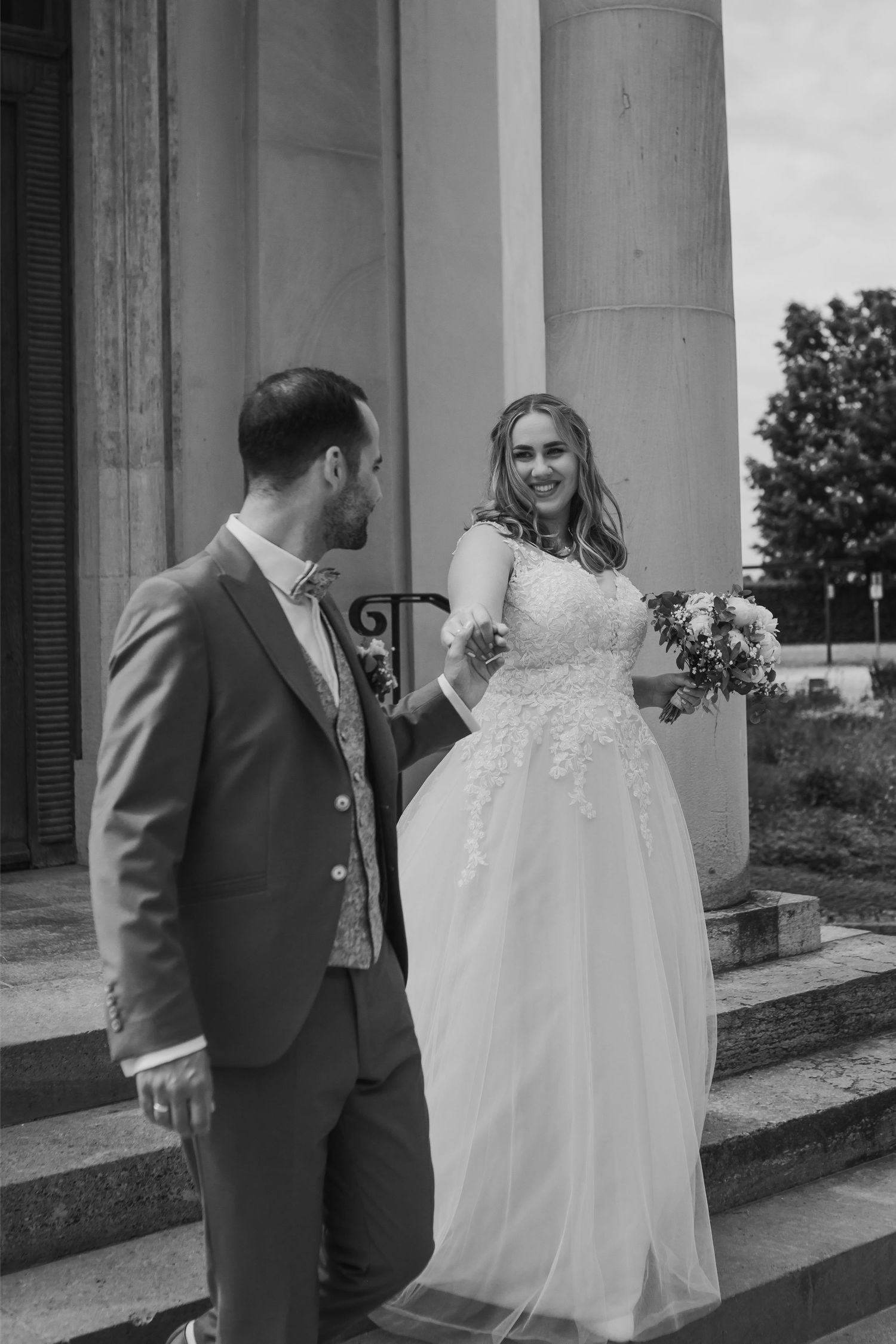 A black and white wedding photo showing a couple on stone steps with classical columns, the bride in a lace gown holding flowers.
