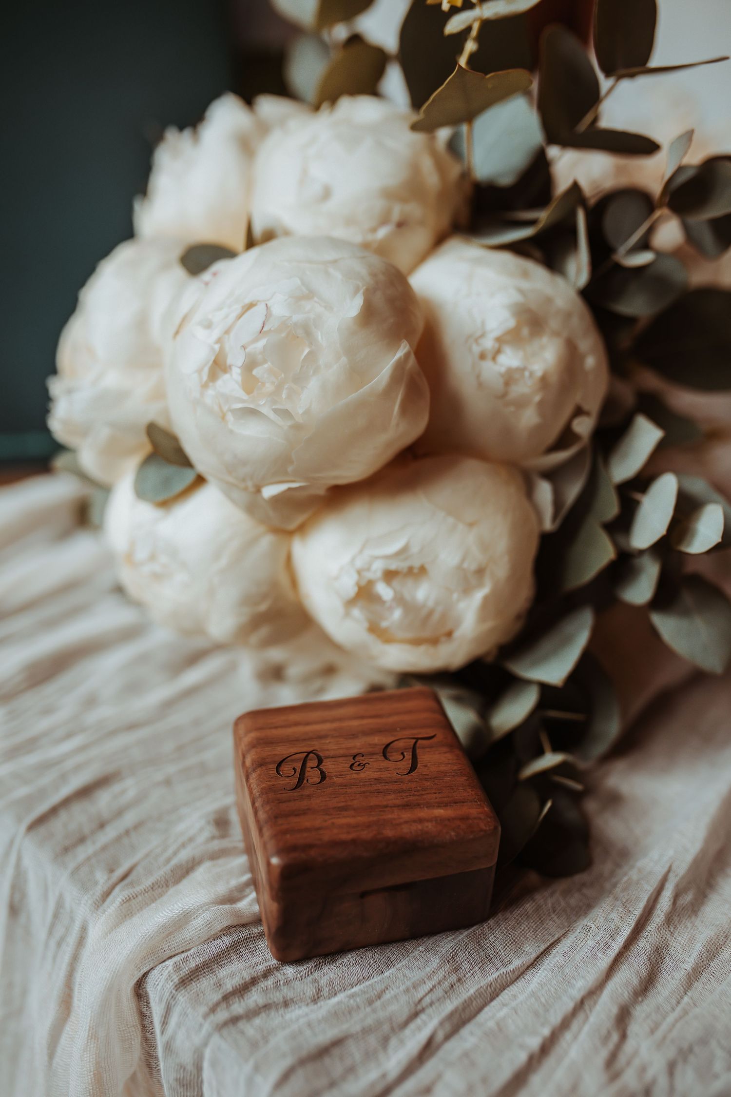 Rustic wooden ring box with monogram beside white peony bouquet on fabric.