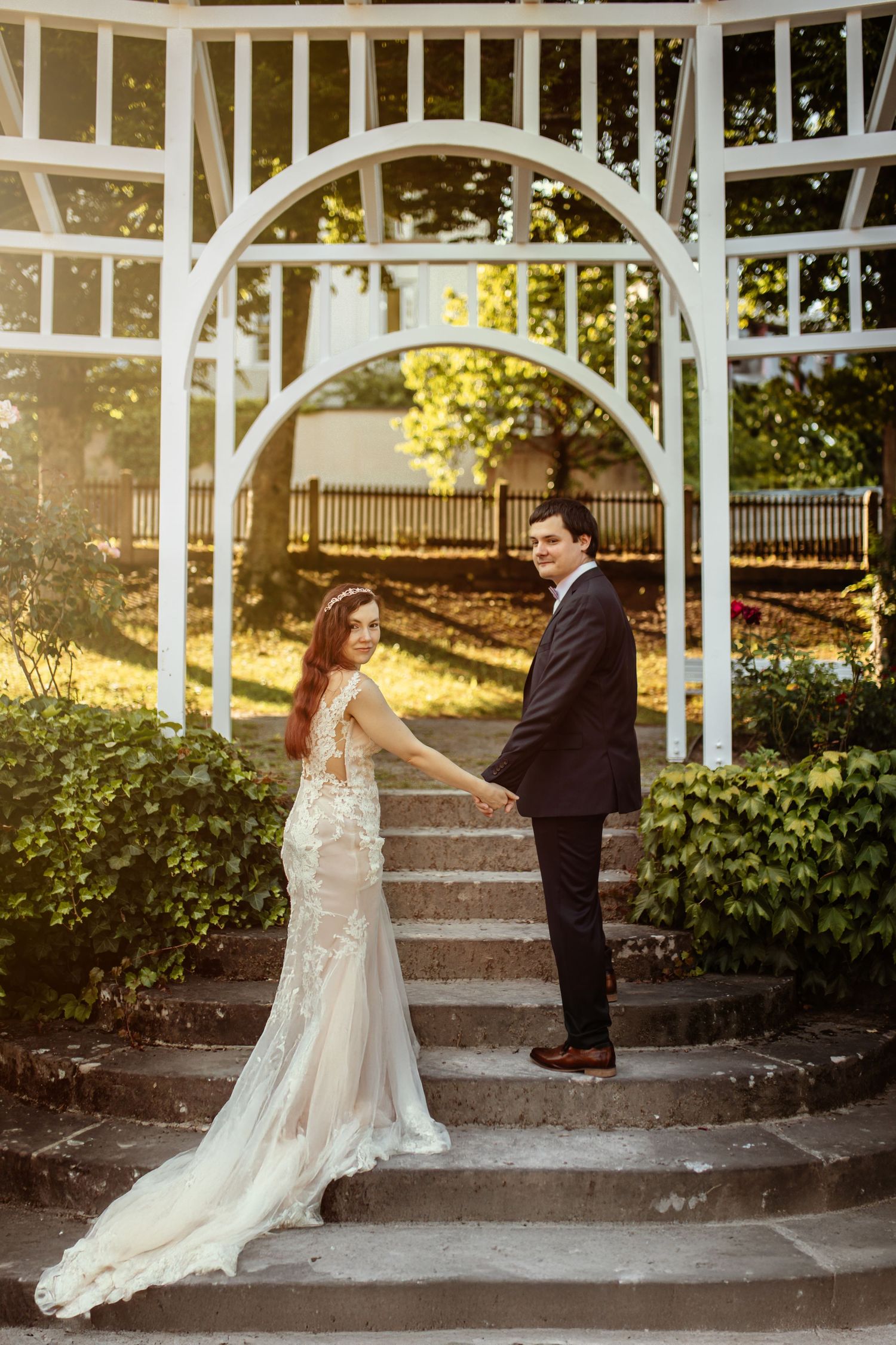 A couple stands on stone steps under a white garden arbor during golden hour, with sunlit foliage in the background.