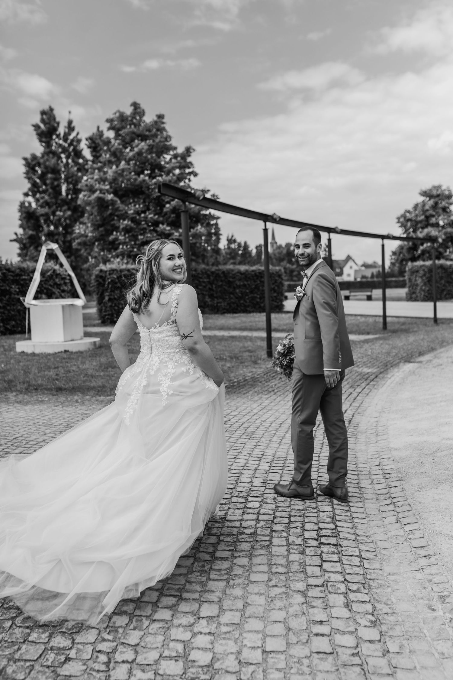 Bride in flowing gown poses playfully while groom stands on cobblestone path.