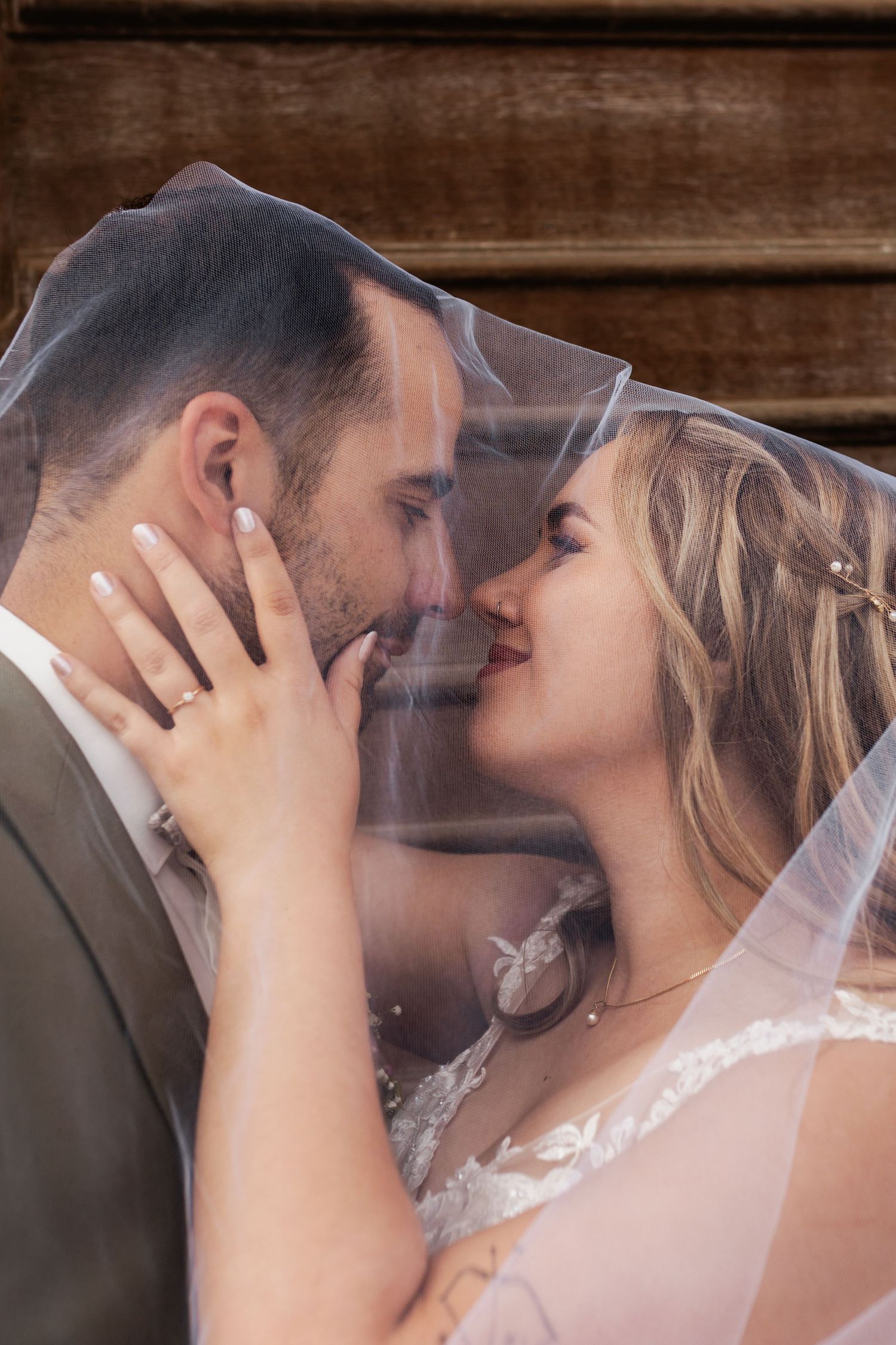 A romantic wedding couple shares an intimate moment under the bride's veil against a rustic wooden background.