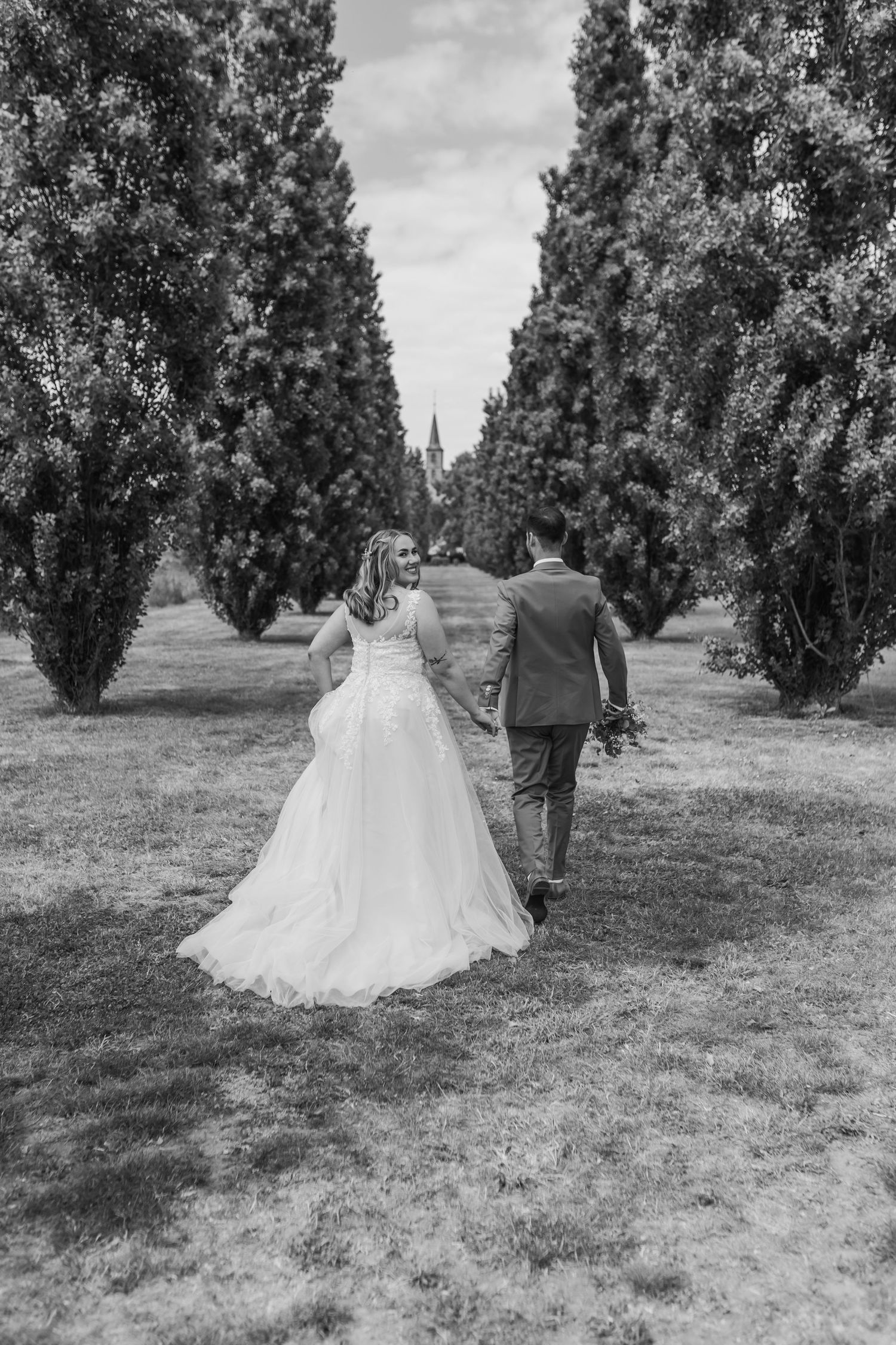 Newlyweds walk hand in hand down a tree-lined path in their wedding attire.