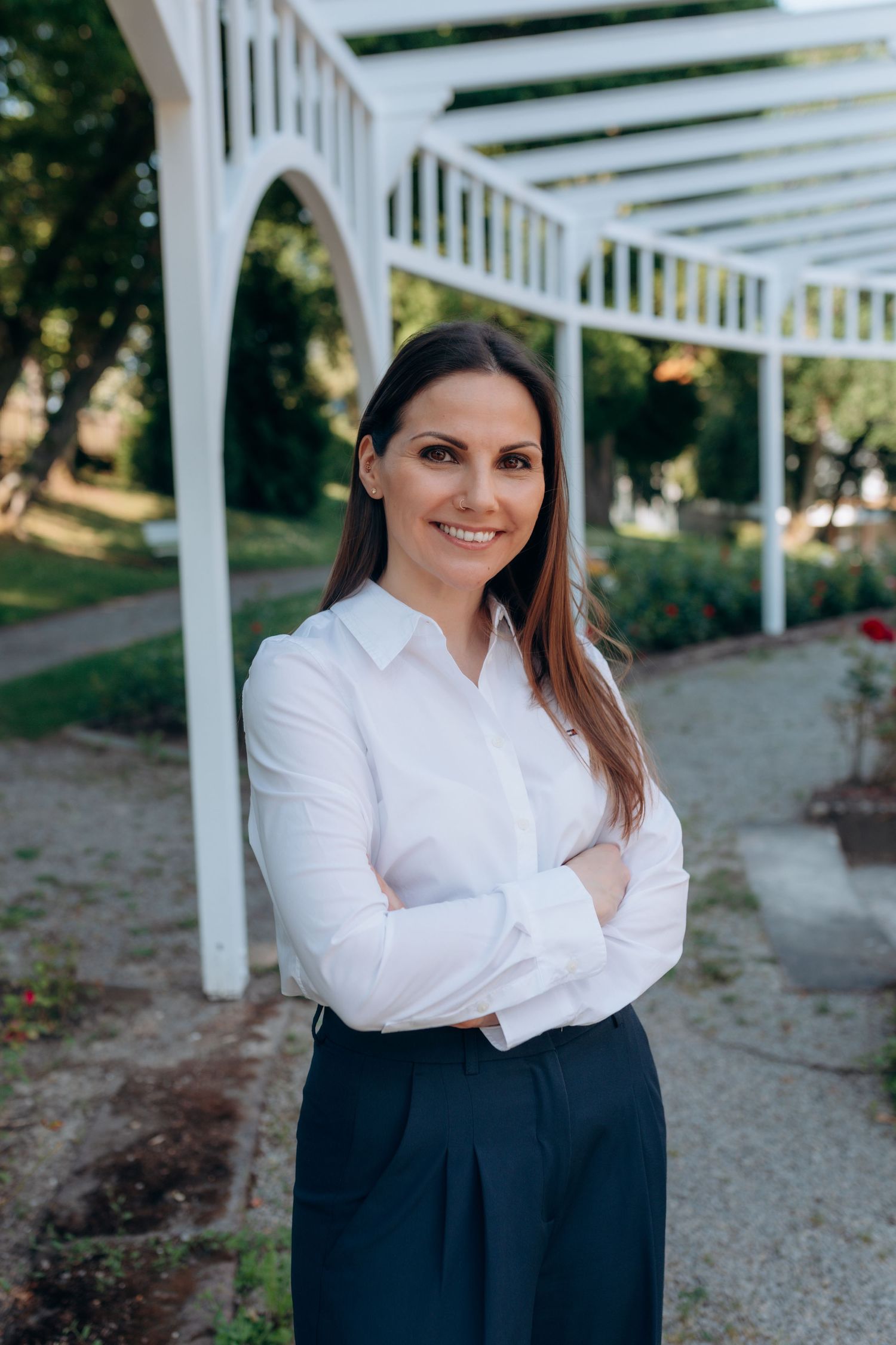 A person in a white button-down shirt and navy skirt stands with crossed arms under a white pergola outdoor structure.