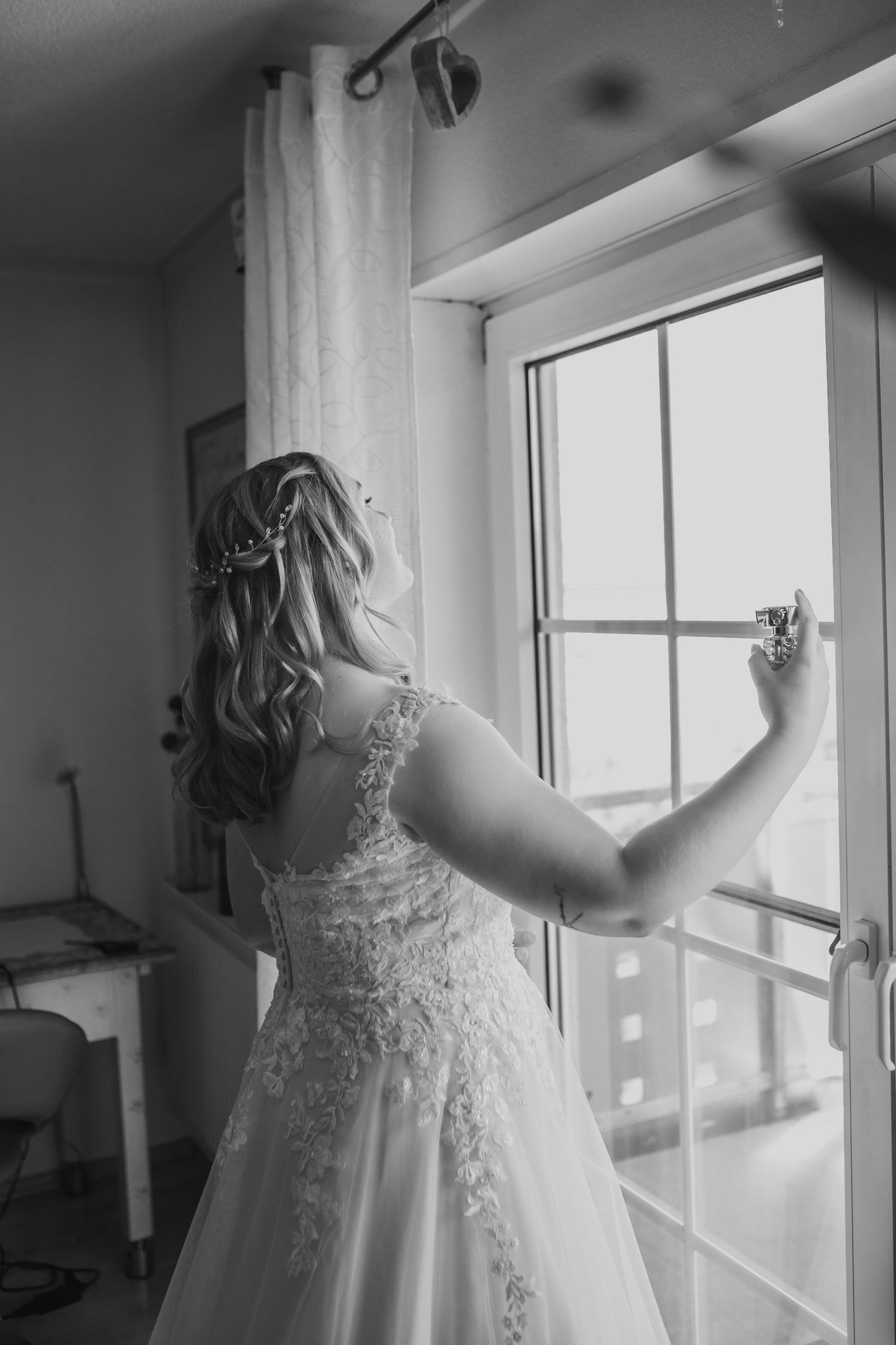 Black and white photo showing back of bride in lace wedding dress looking out window in romantic pose.