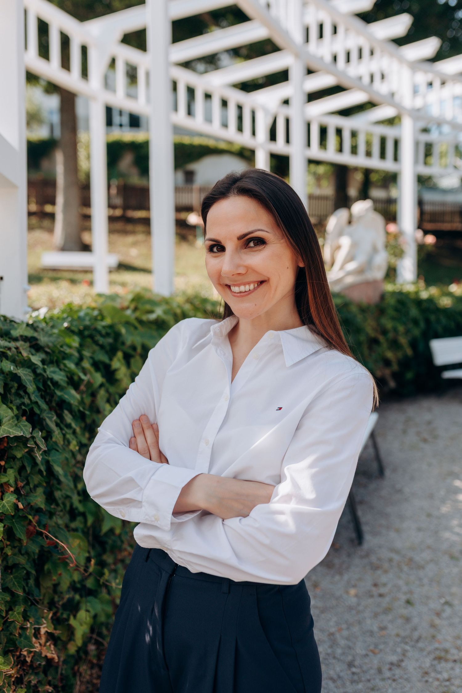 Professional in crisp white shirt and dark skirt stands confidently with arms crossed in outdoor garden setting.
