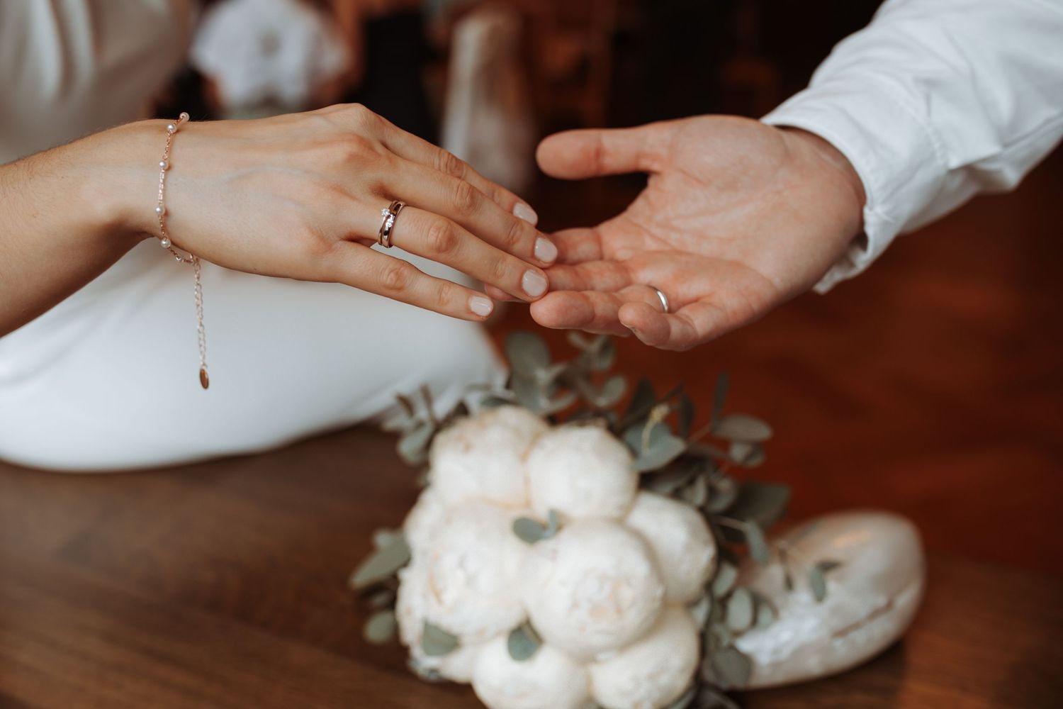Close-up of wedding rings and hands with white peony bouquet on wooden surface.