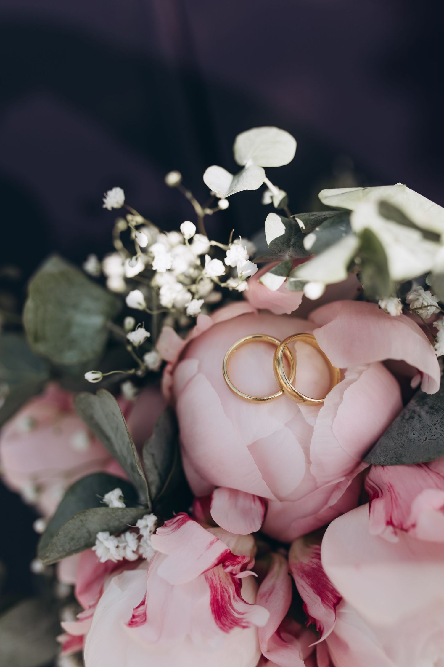 Gold wedding rings nestled on pink peony flower with baby's breath and eucalyptus leaves in a romantic arrangement.