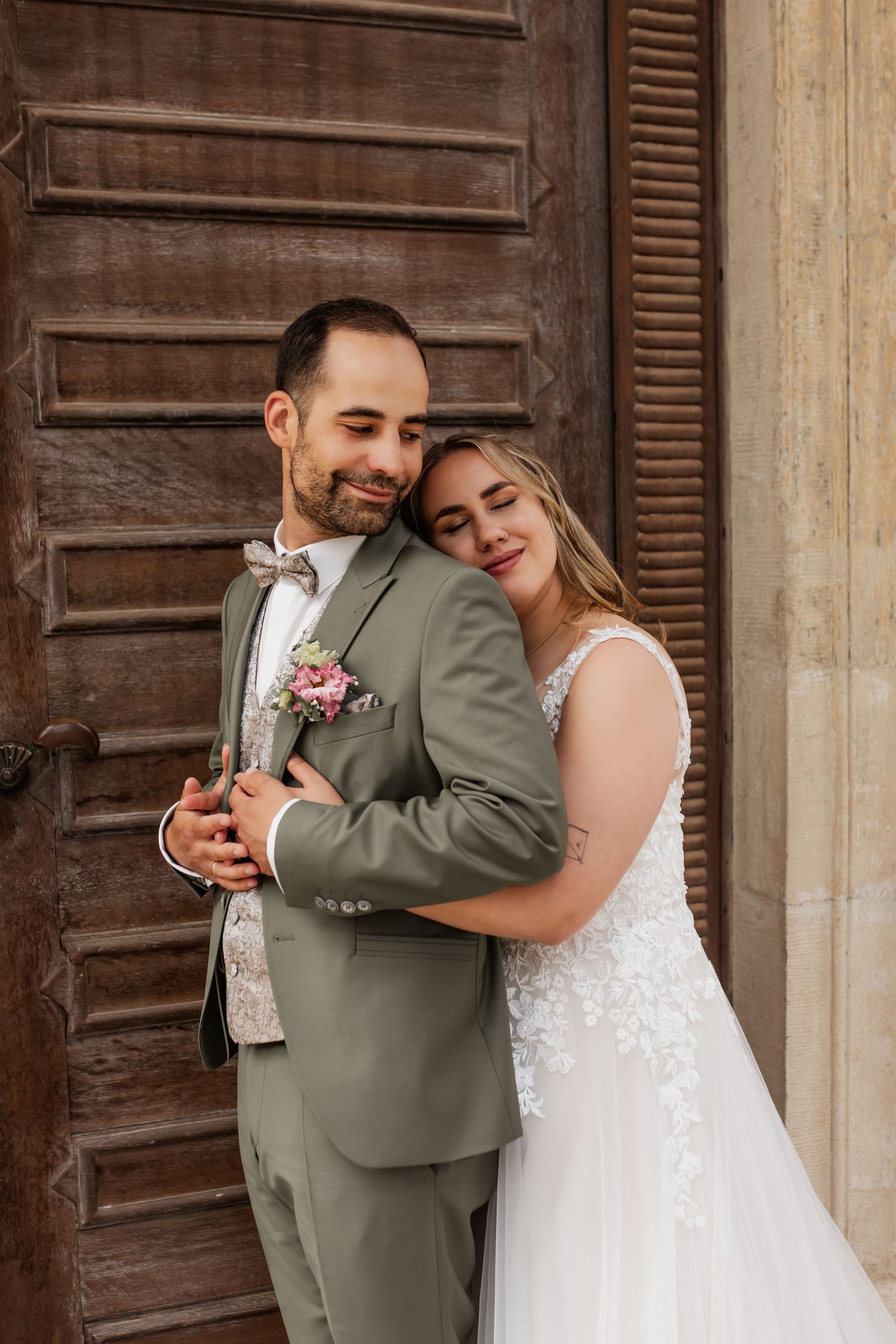 A romantic wedding couple embraces against a rustic wooden door, with the groom in sage green suit and pink boutonniere.