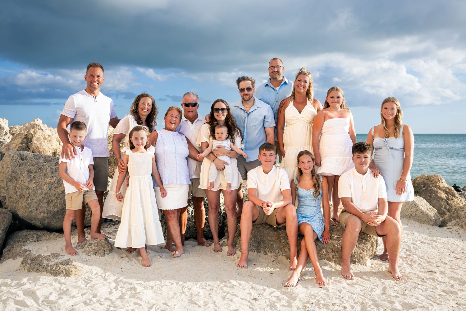 Large family group photo on a sandy beach with rocky backdrop and blue ocean, all wearing white and khaki outfits.