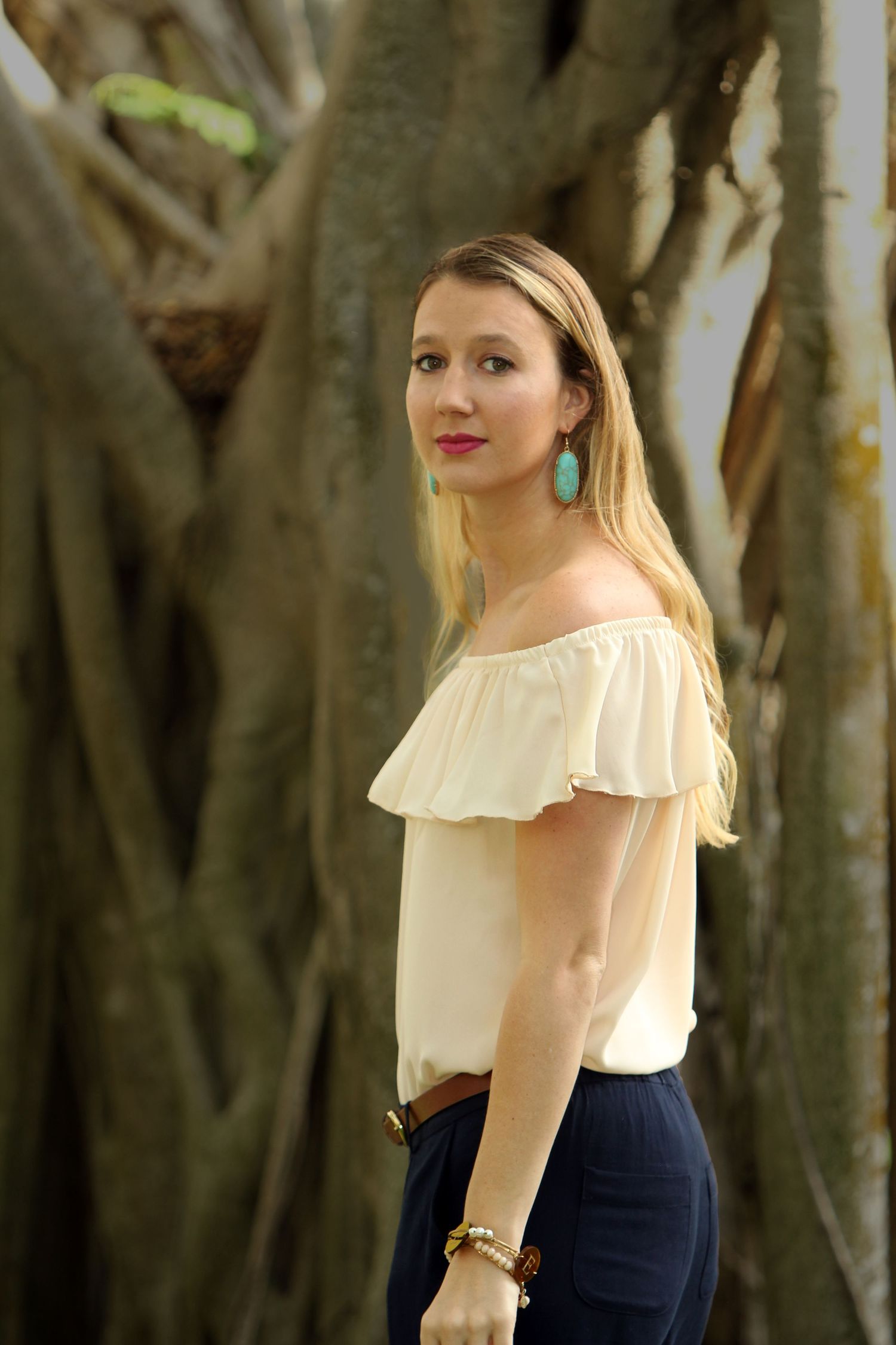 Portrait shot against banyan trees showing off-shoulder white top and turquoise earrings.