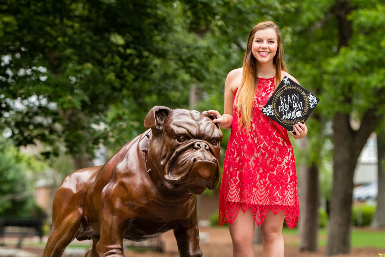 Student in red lace dress poses with bronze bulldog statue on campus.