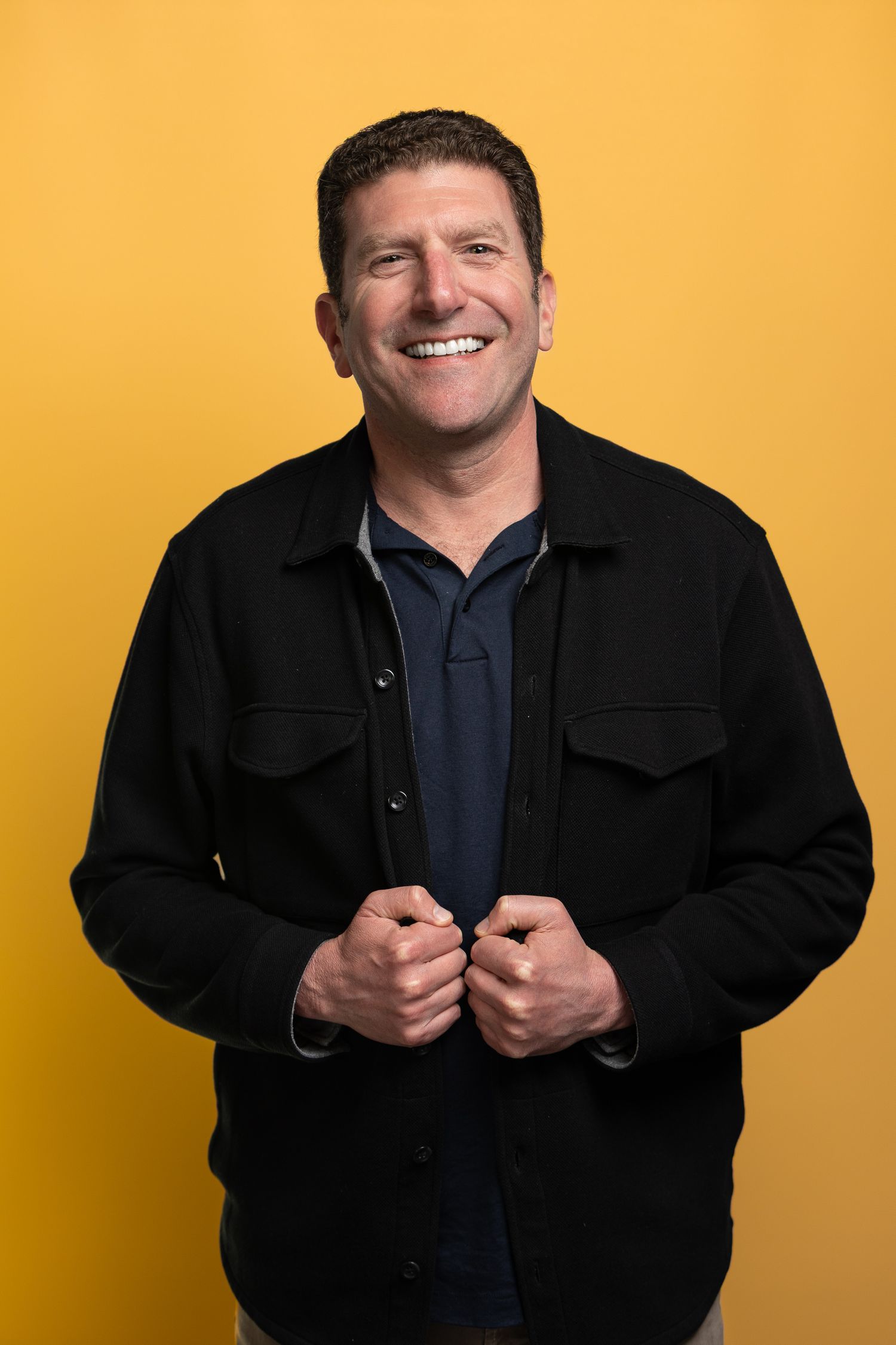 Smiling middle-aged man in black jacket poses confidently against a bright yellow background.