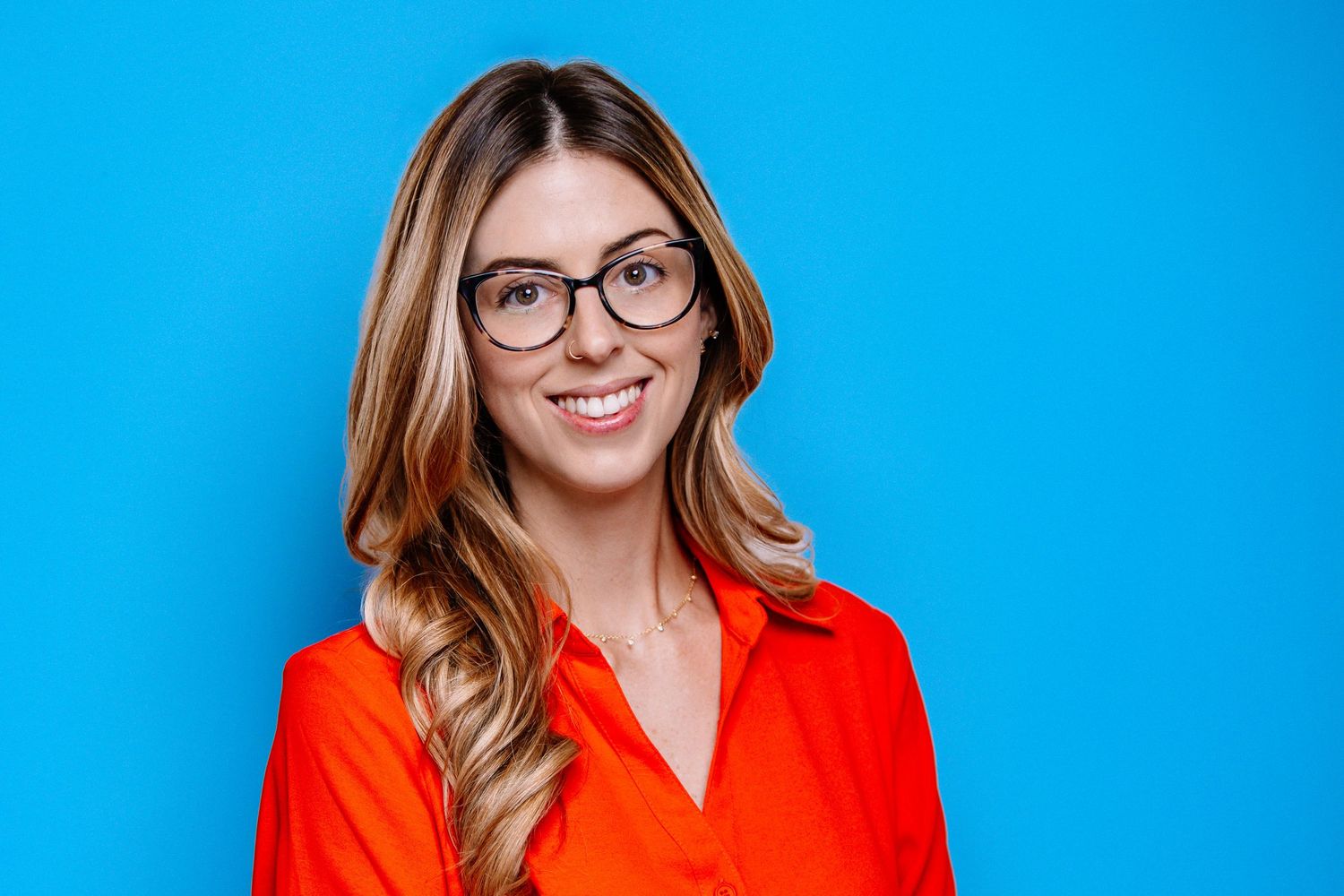 Professional headshot of someone wearing glasses and a bright red blazer against a vibrant blue background.