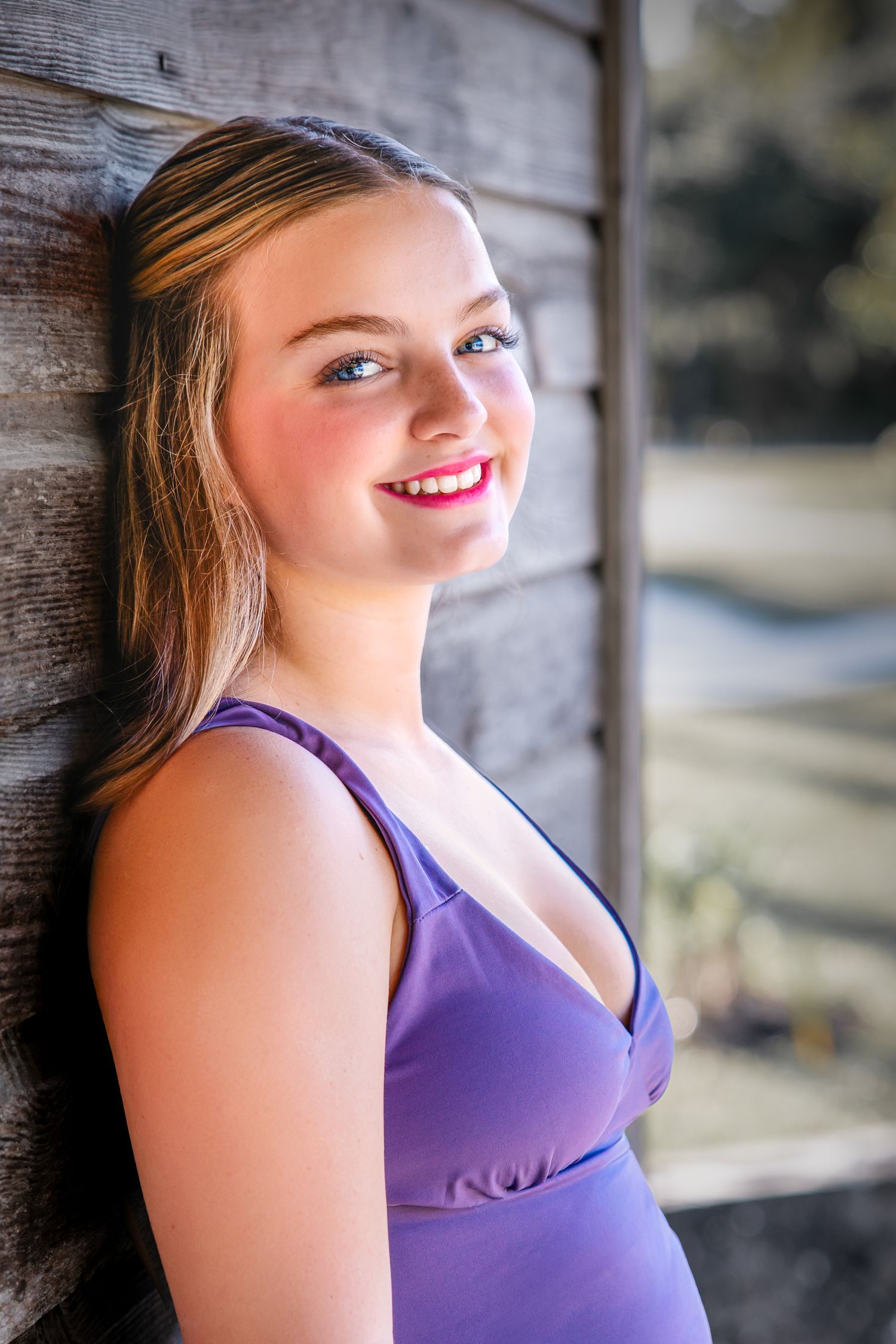 A portrait of someone in a purple tank top leaning against a wooden wall while smiling in natural lighting.