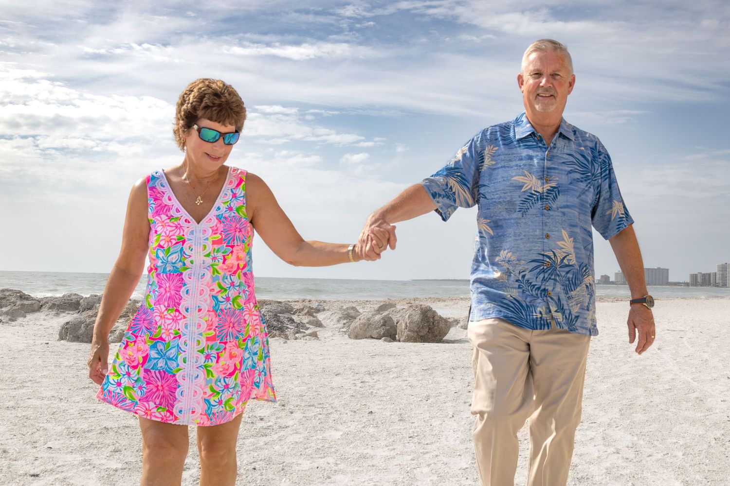 A couple walks hand in hand along a sunny white sand beach wearing colorful summer attire.