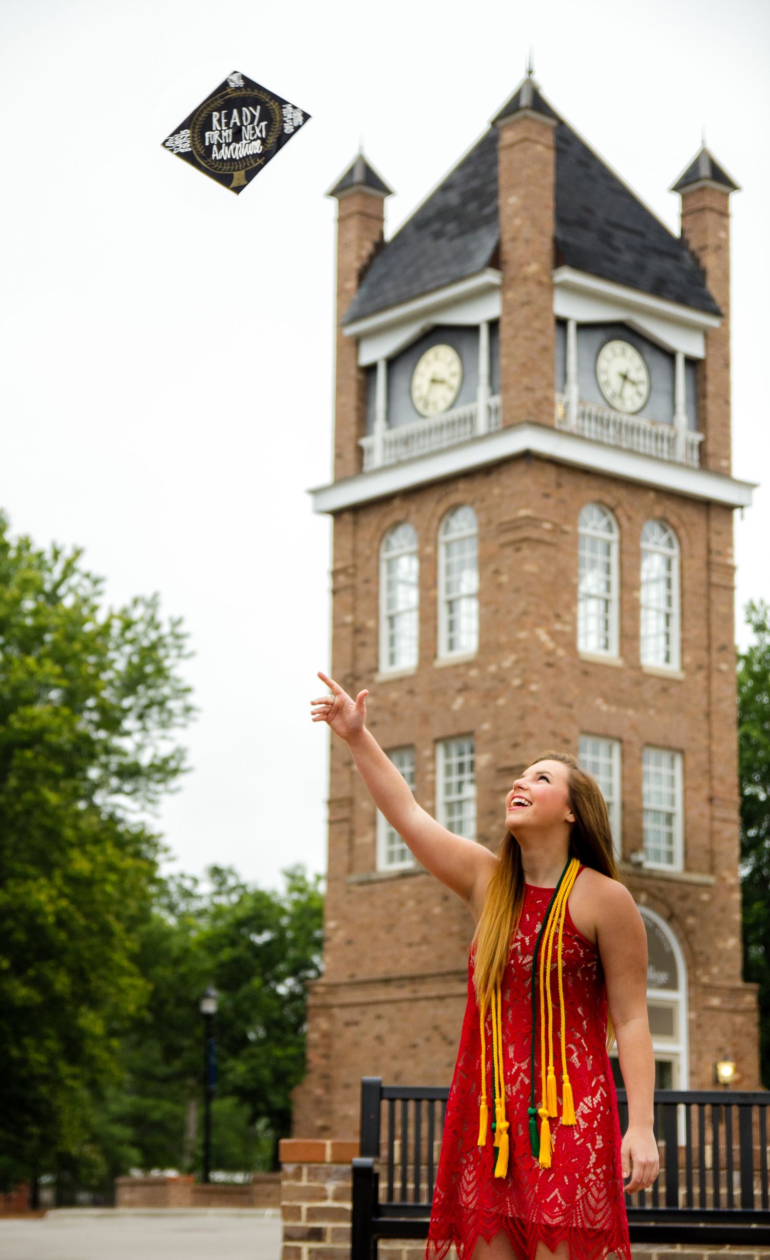 A graduate in red and gold regalia celebrates joyfully in front of a brick clock tower building on campus.