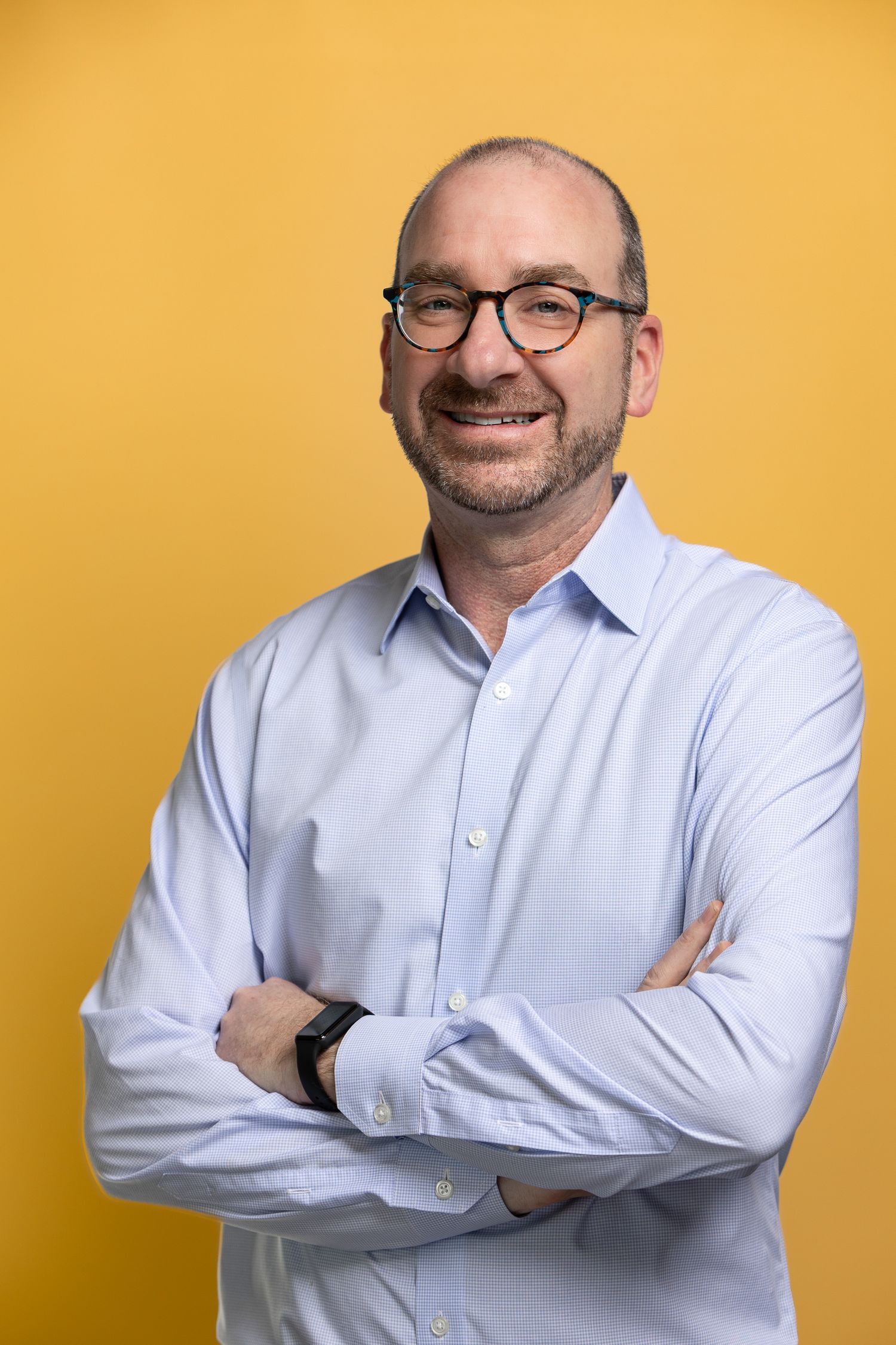 Middle-aged man with glasses and stubble smiles confidently, arms crossed, wearing a light blue shirt on yellow background.