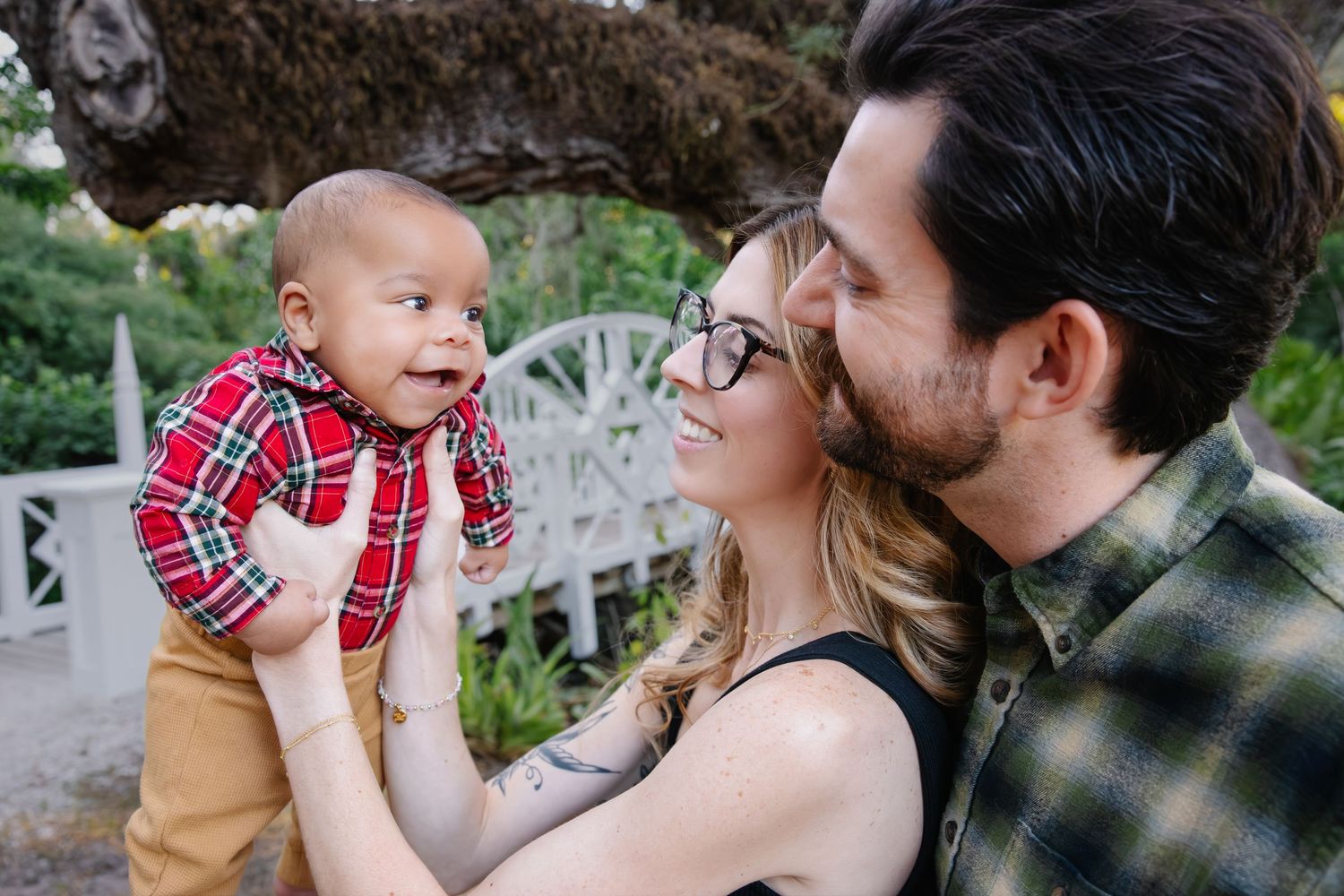 A family shares a tender outdoor moment together on a garden patio with their baby wearing a red plaid shirt.