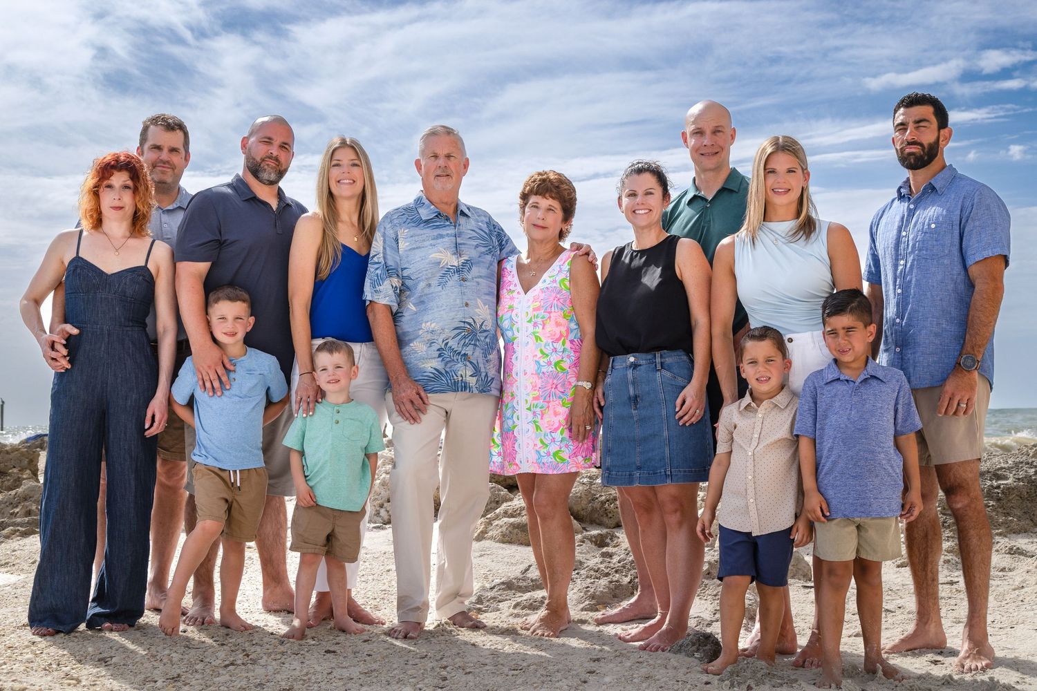 A large family group poses together on a sunny beach with blue sky and white clouds in the background.