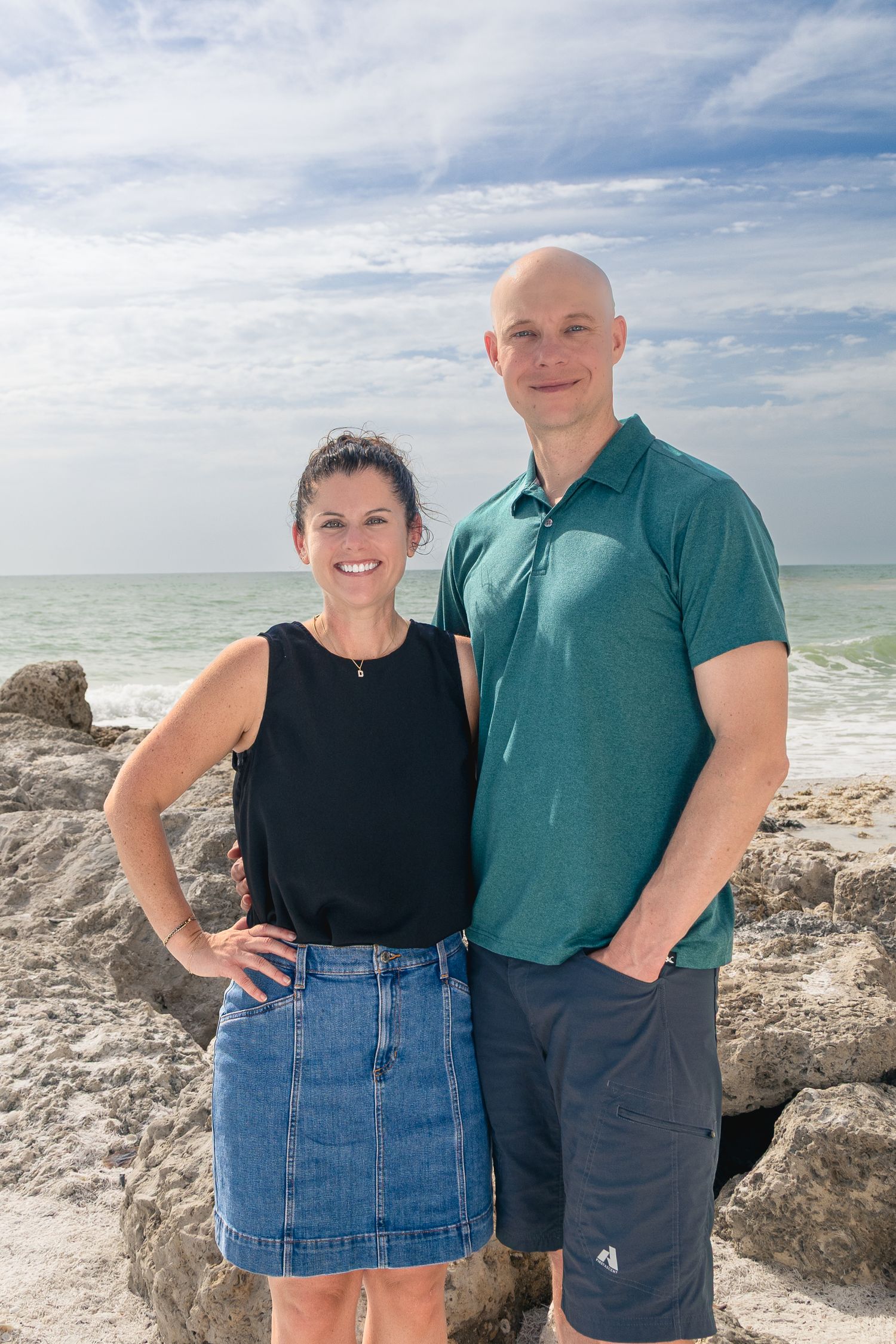 A couple stands together on a rocky beach with blue ocean waves and cloudy sky in the background.