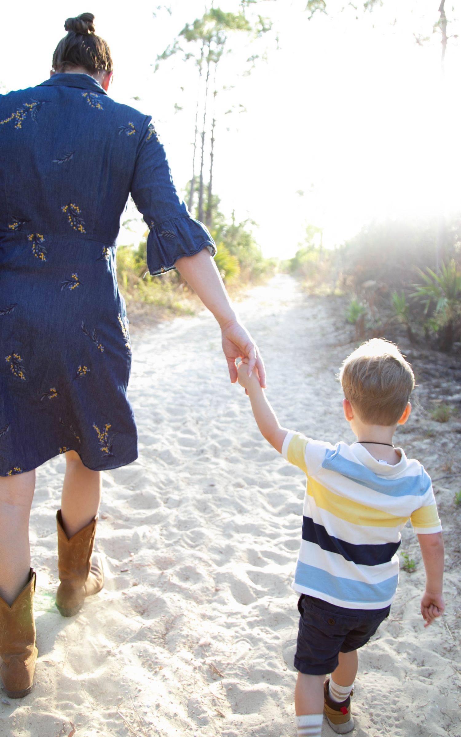 Person in blue dress holding hands with small child while walking on sandy path.