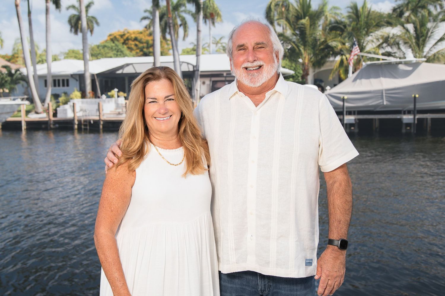 Two people in white attire smile while standing at a marina with boats and palm trees in the background.