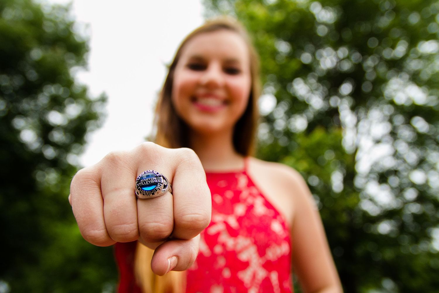 A close-up view of a championship ring being worn on a hand with a red dress visible in the background.