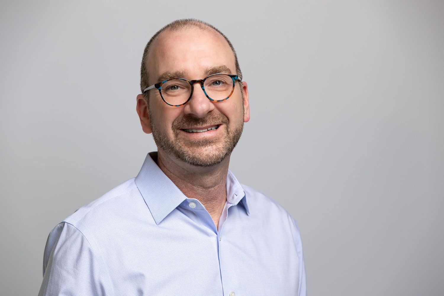 Professional headshot of a bald man with glasses wearing a light blue shirt, smiling against a gray background.