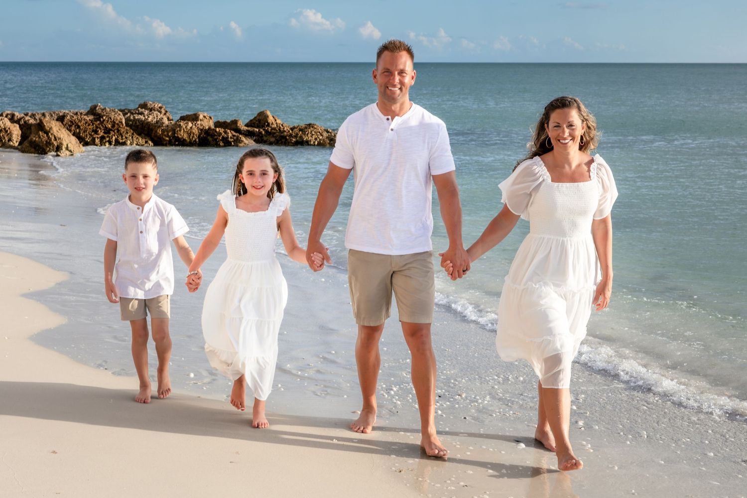 Family of four holding hands walking on a sunny beach, wearing white outfits with ocean waves in the background.