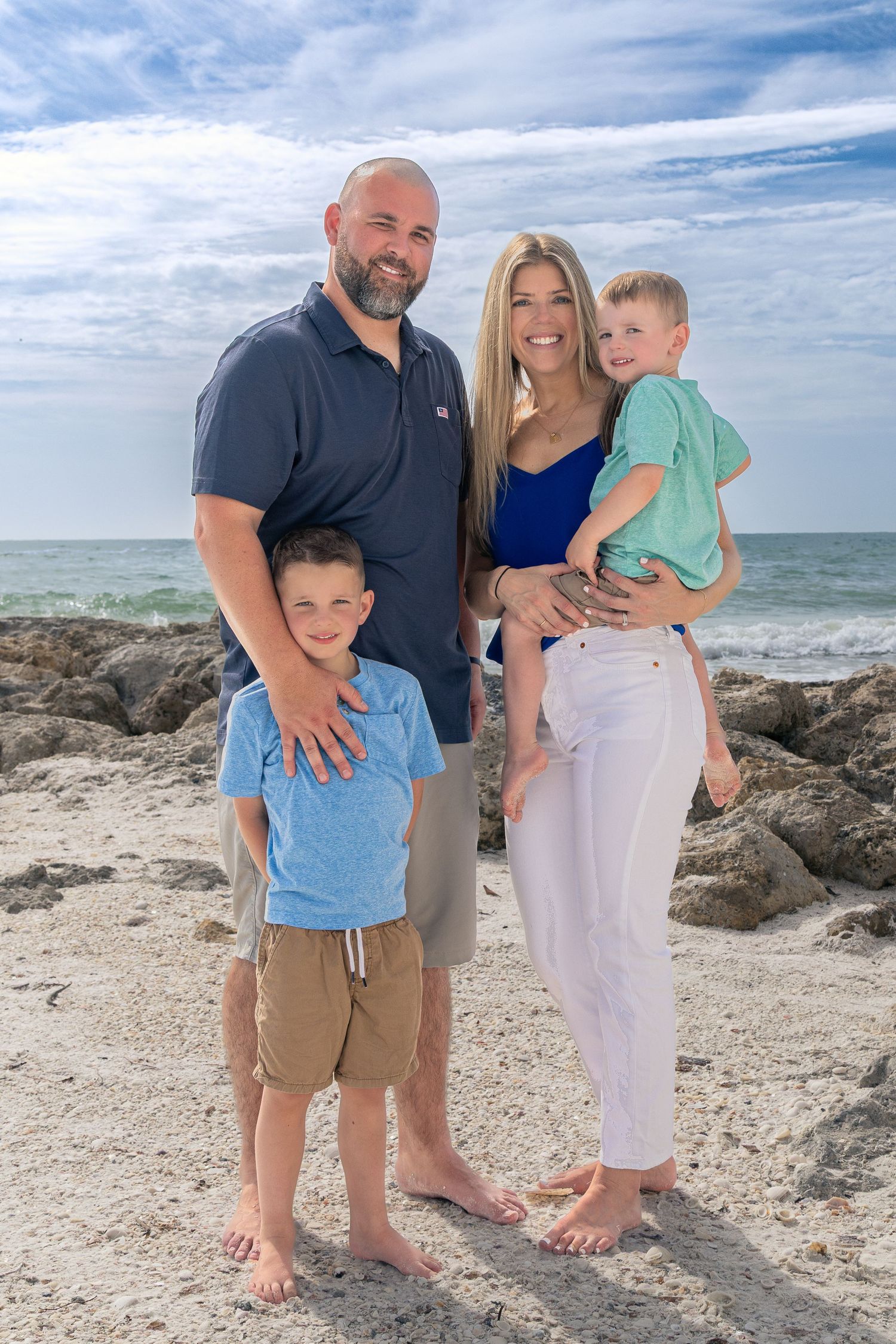 A family poses together on a sunny beach with rocky coastline and ocean waves in the background.