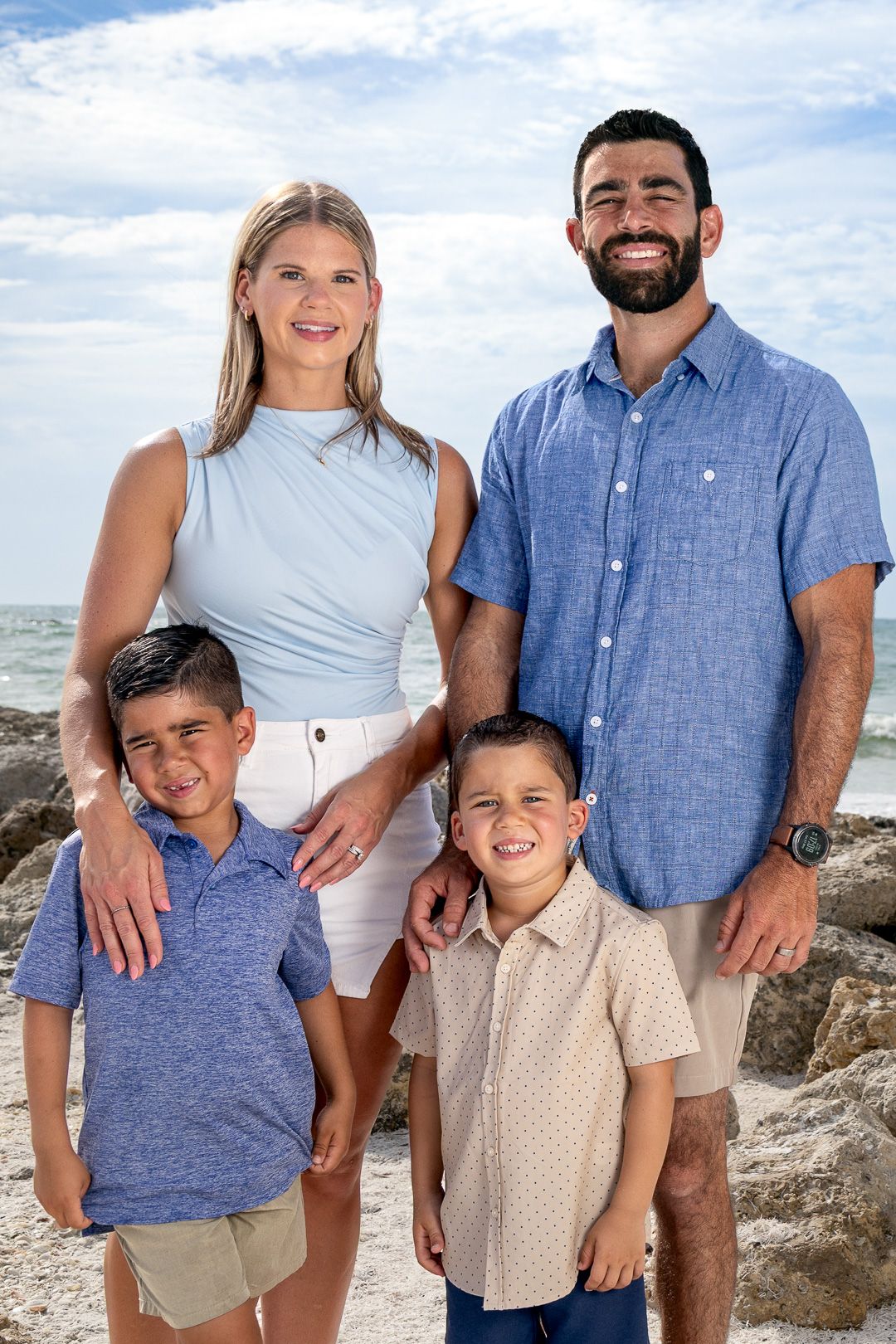 Family wearing coordinated blue and white outfits poses together on rocky beach landscape.