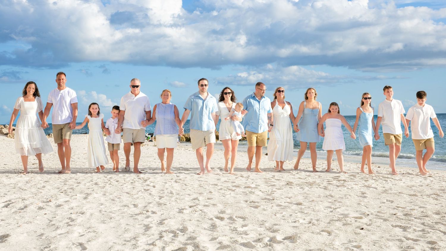 Large family group posing together on a sunny white sand beach, dressed in coordinating white and blue outfits.