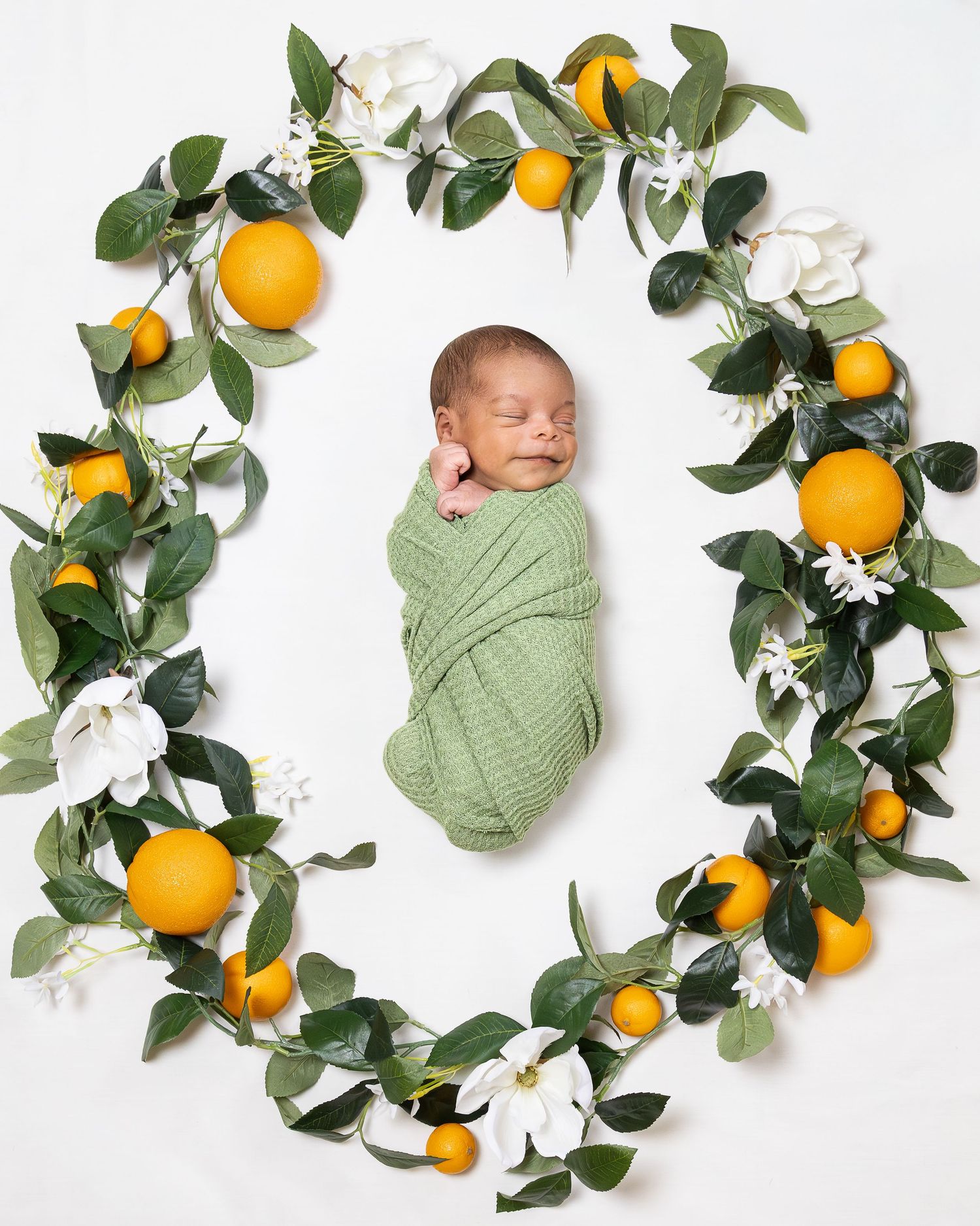 A newborn wrapped in sage green swaddle lies surrounded by fresh oranges, greenery, and white flowers in a circular arrangement.