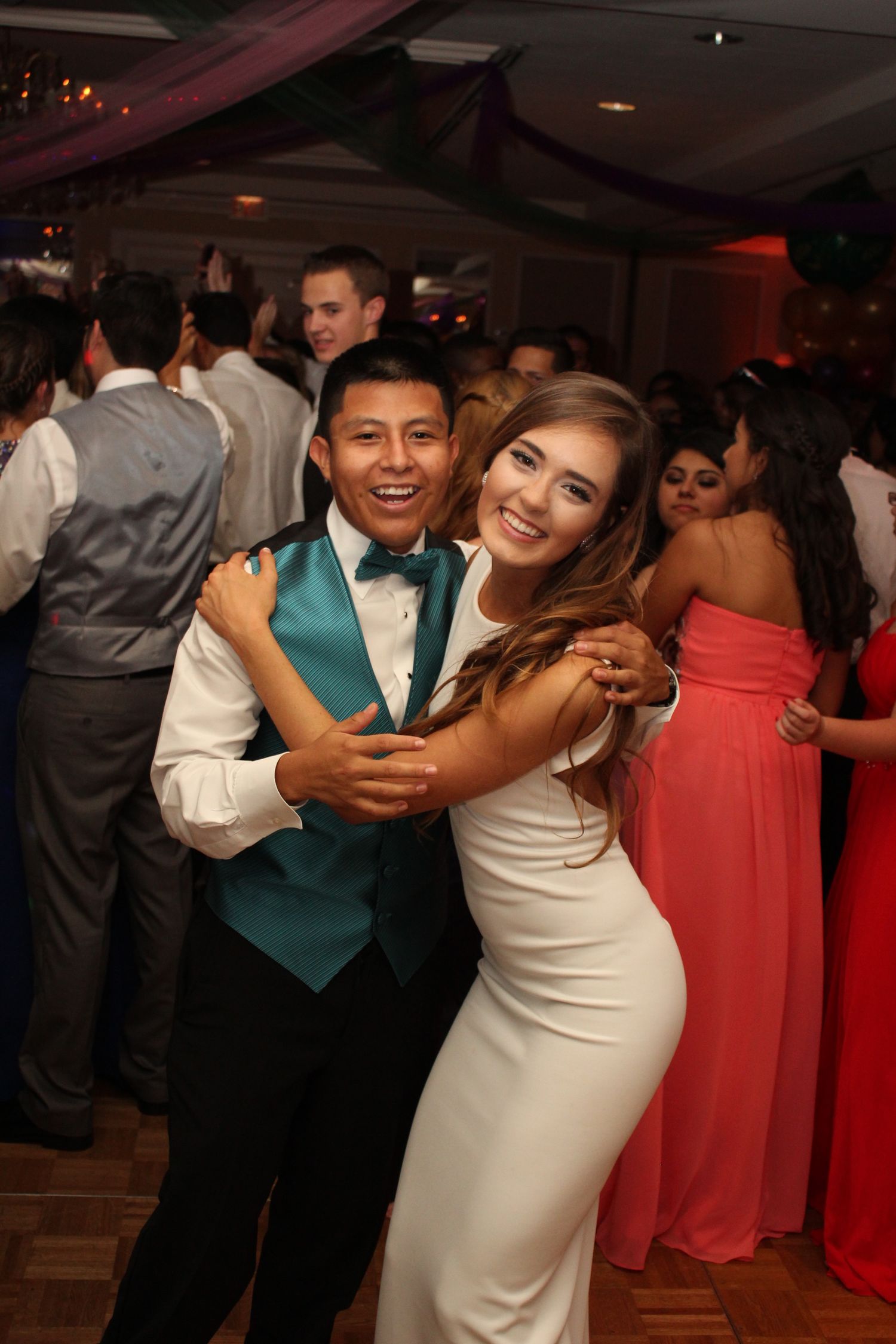 Couple dancing and smiling at formal event with guest in teal vest and white dress.