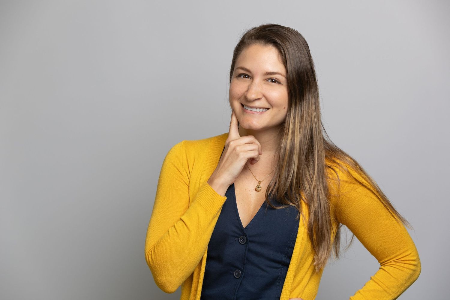 Smiling young woman in yellow cardigan posing confidently with hand on chin against gray background.