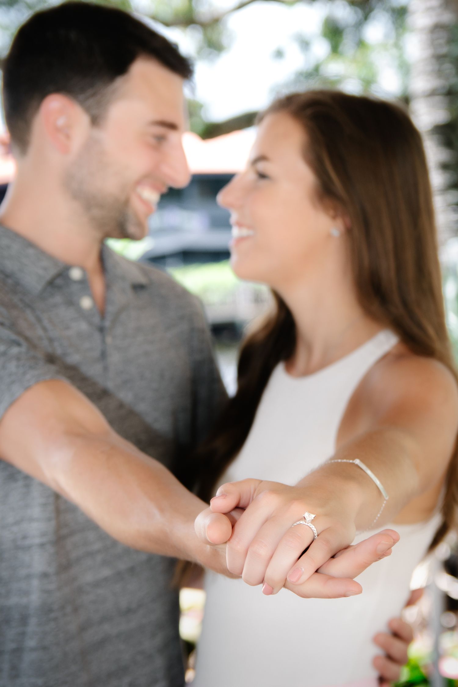 A couple shares a romantic moment while holding hands and displaying an engagement ring outdoors.