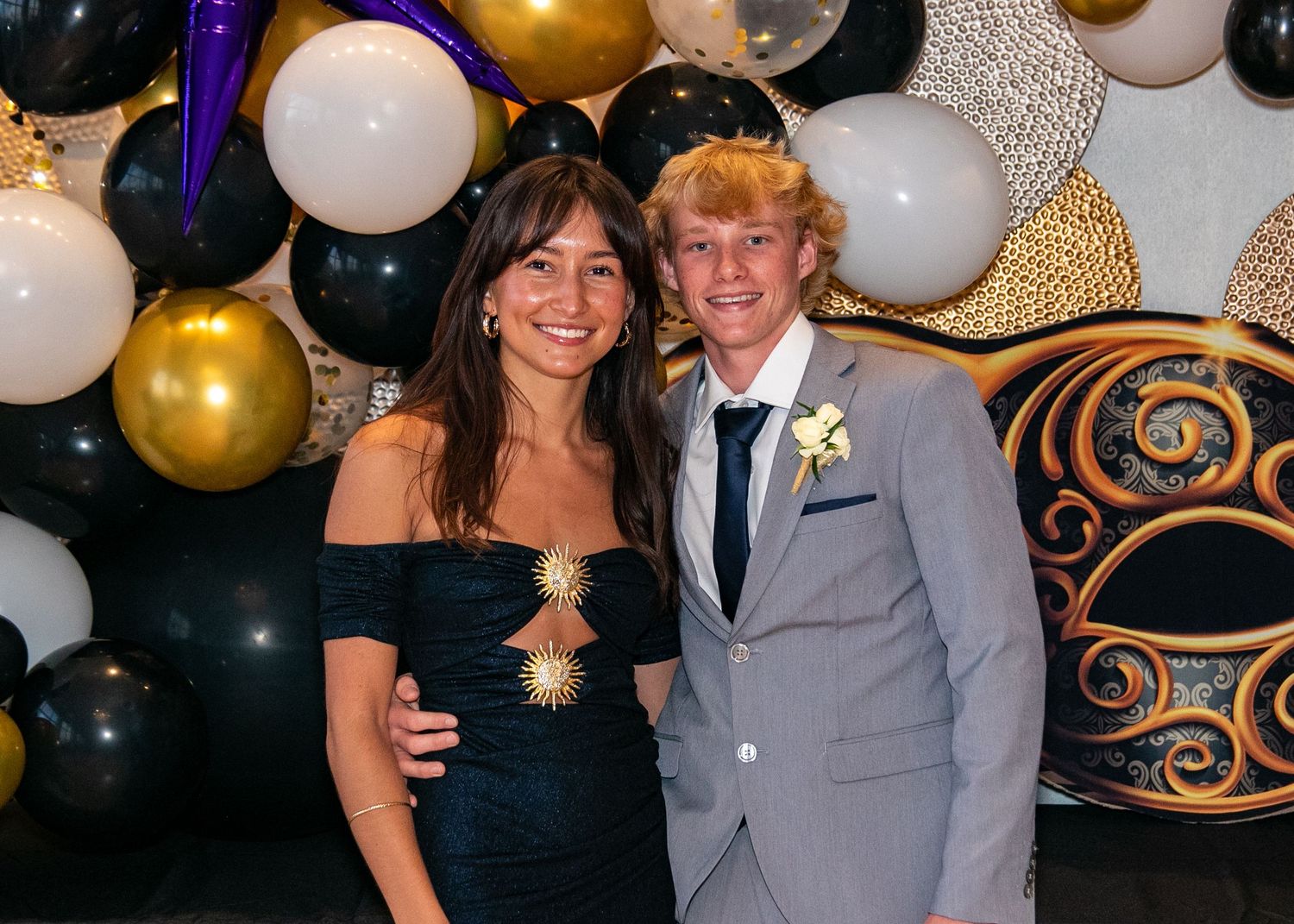 Couple poses in front of black and gold balloon display at formal event.