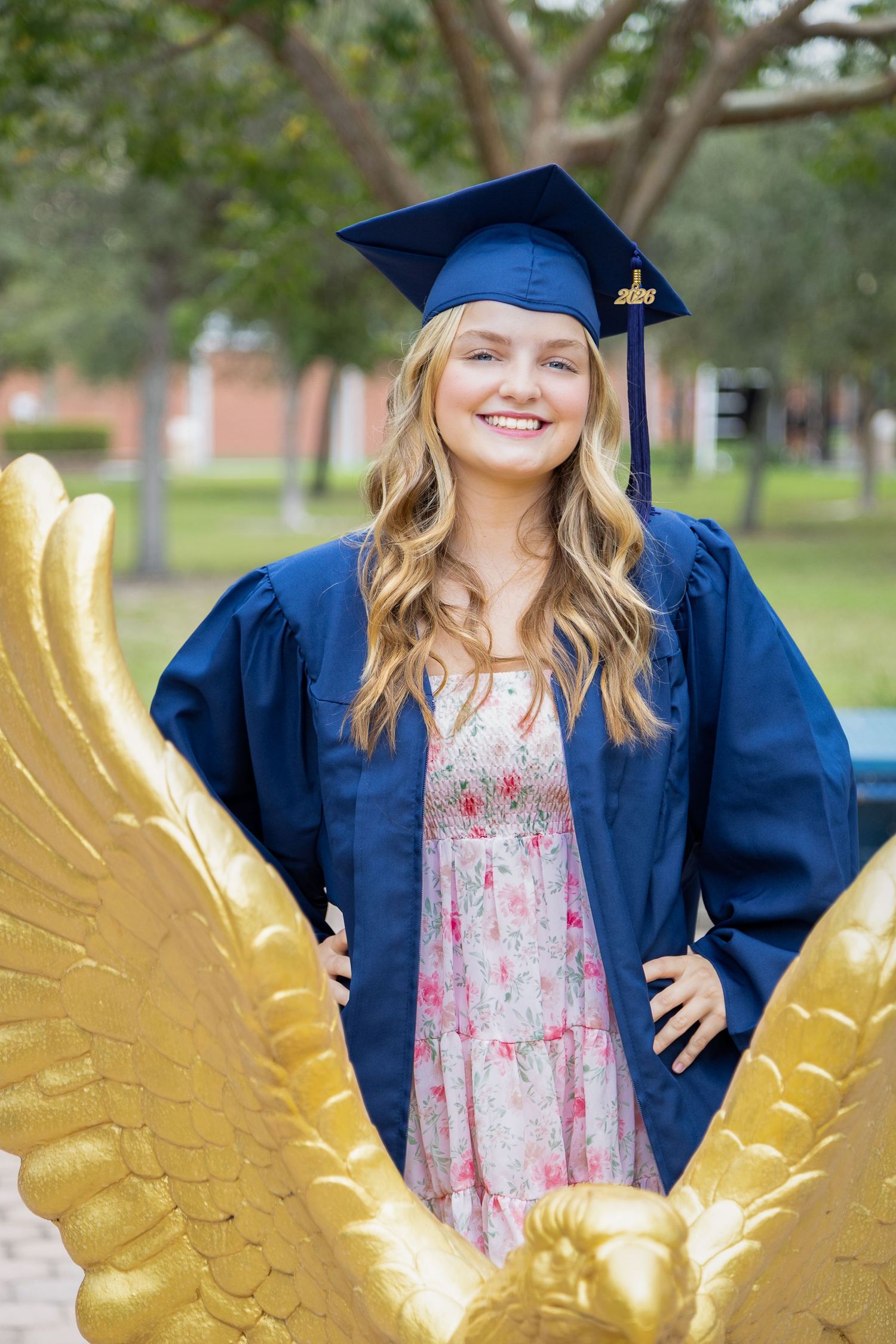 Graduate in blue cap and gown poses with decorative golden wings in an outdoor setting.