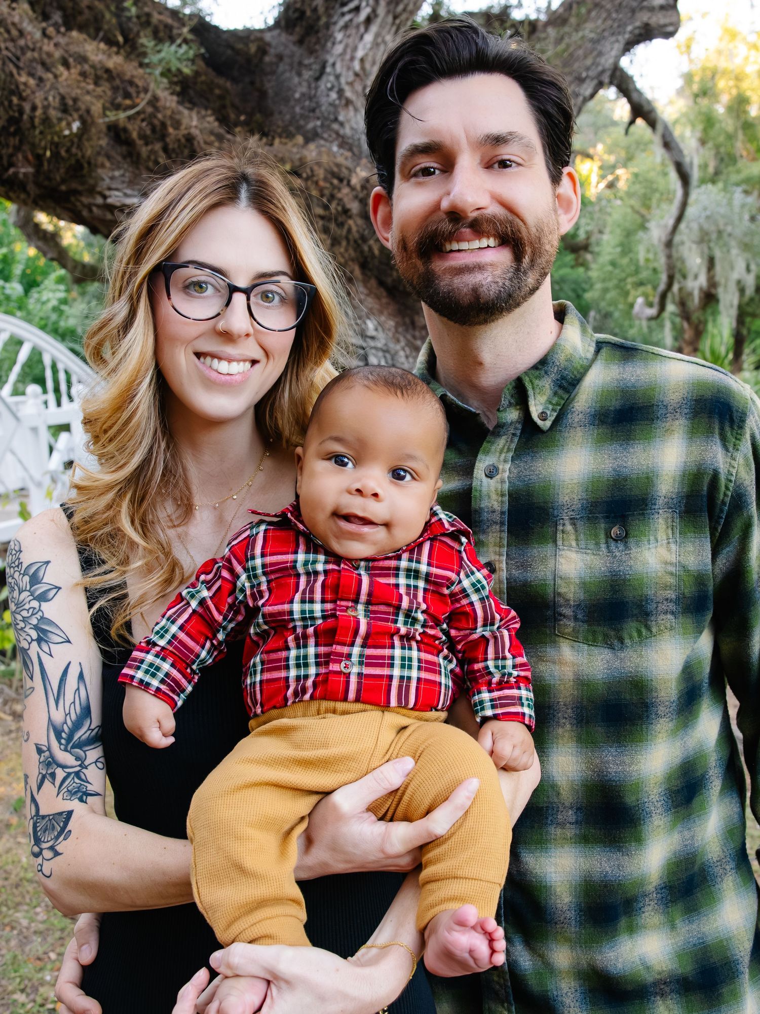 Family wearing coordinating plaid and black outfits poses together outdoors in a natural setting during autumn.