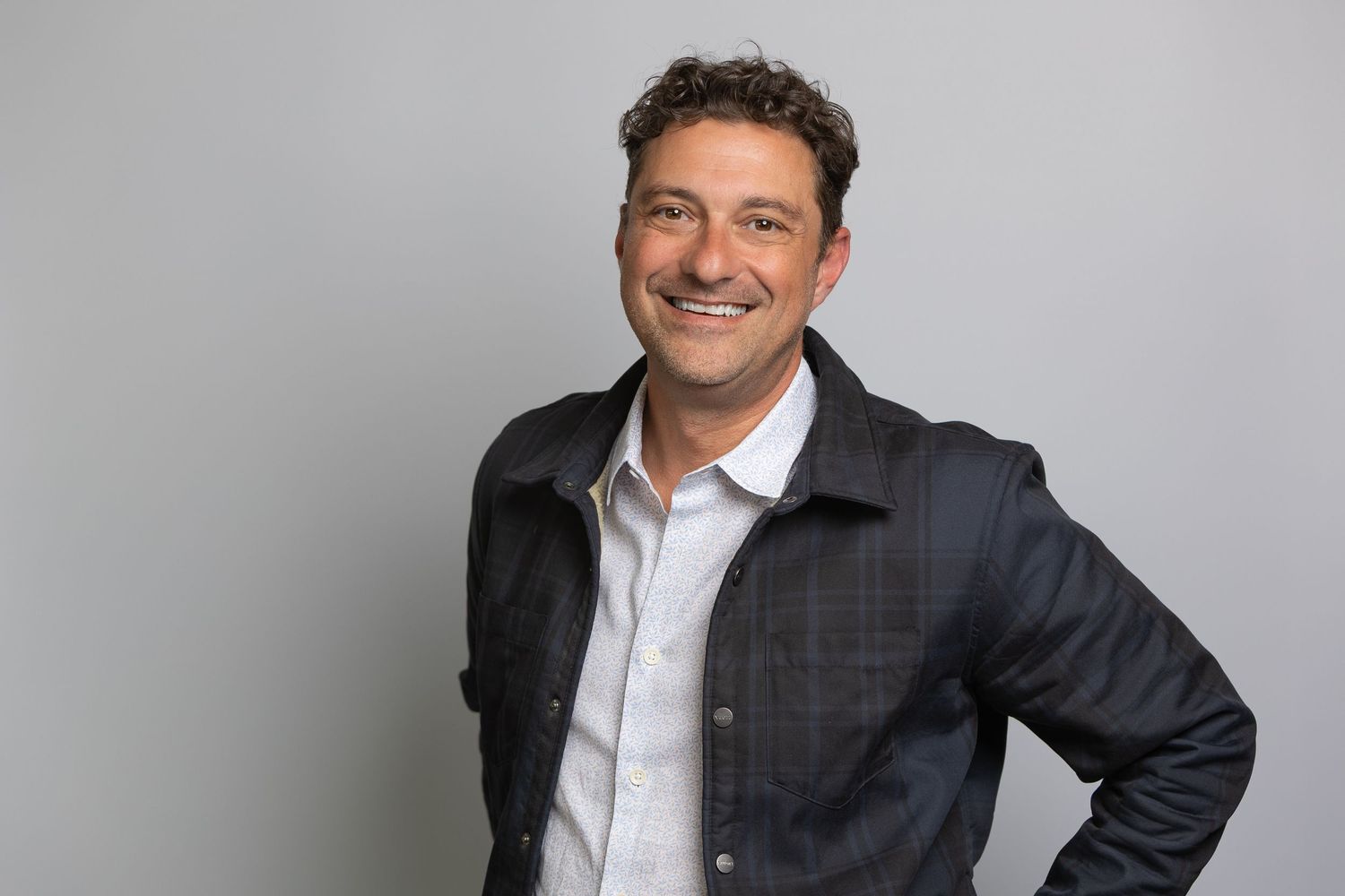 Smiling man in dark jacket and white shirt poses confidently against gray background in professional headshot.