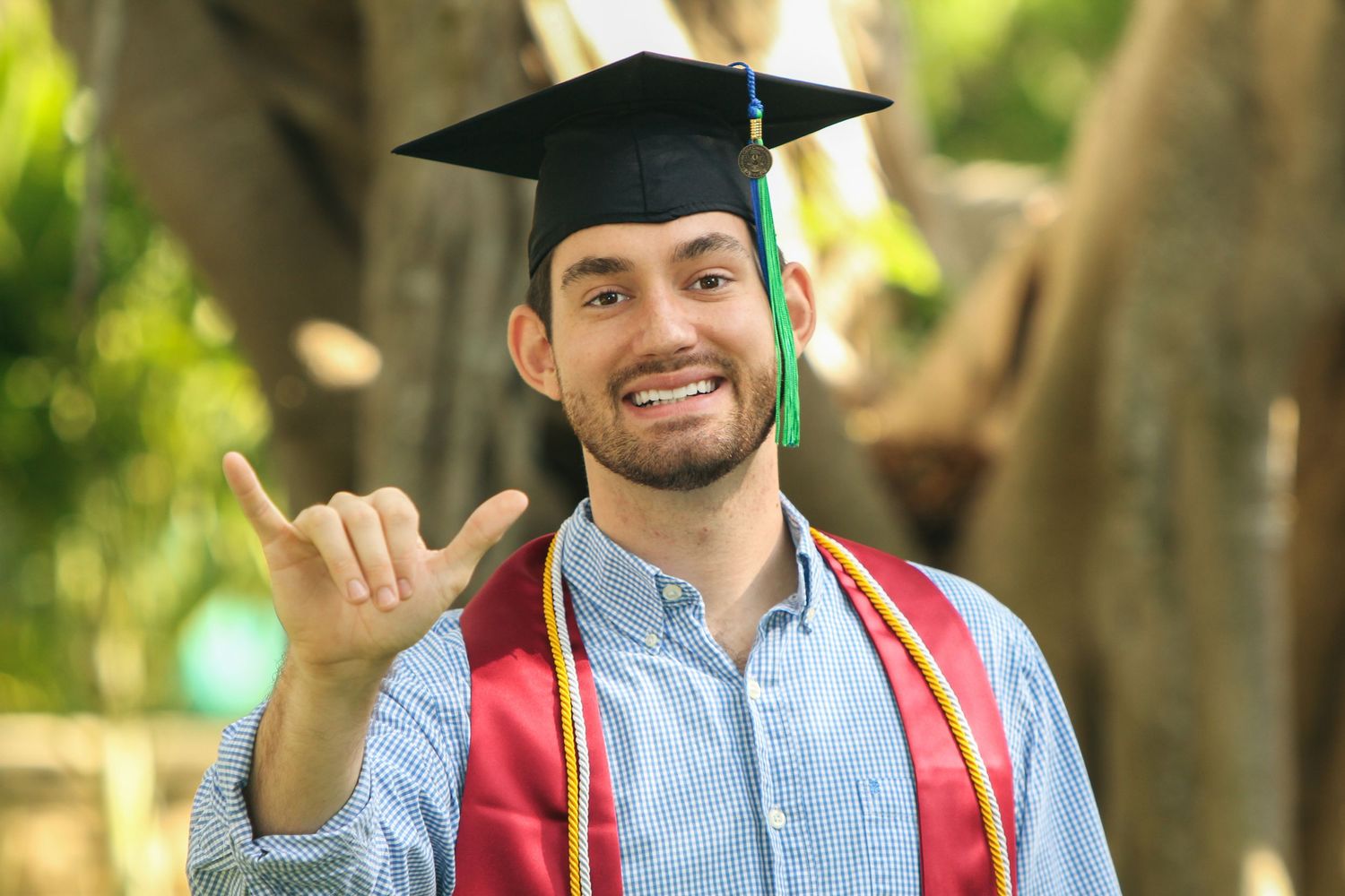 Graduate in cap and gown smiling and gesturing with hand while standing outdoors in natural lighting.