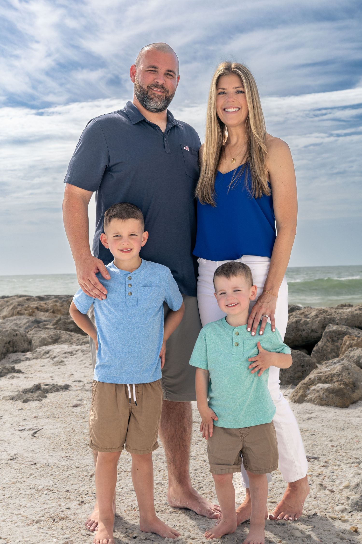 Family of four poses together on a rocky beach with ocean waves and cloudy sky in the background.