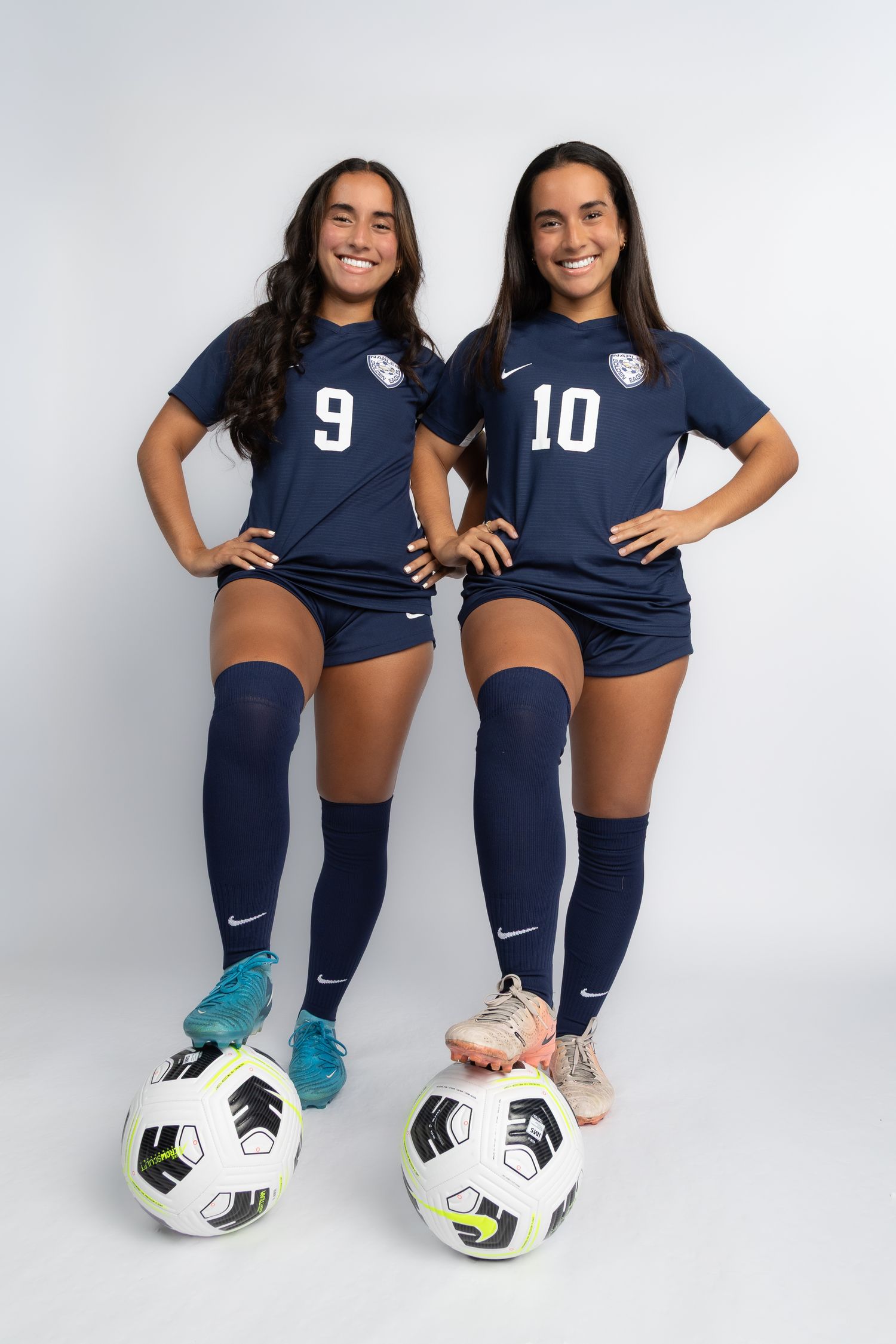 Two soccer players in navy blue uniforms with numbers 9 and 10 pose with soccer balls against white background.