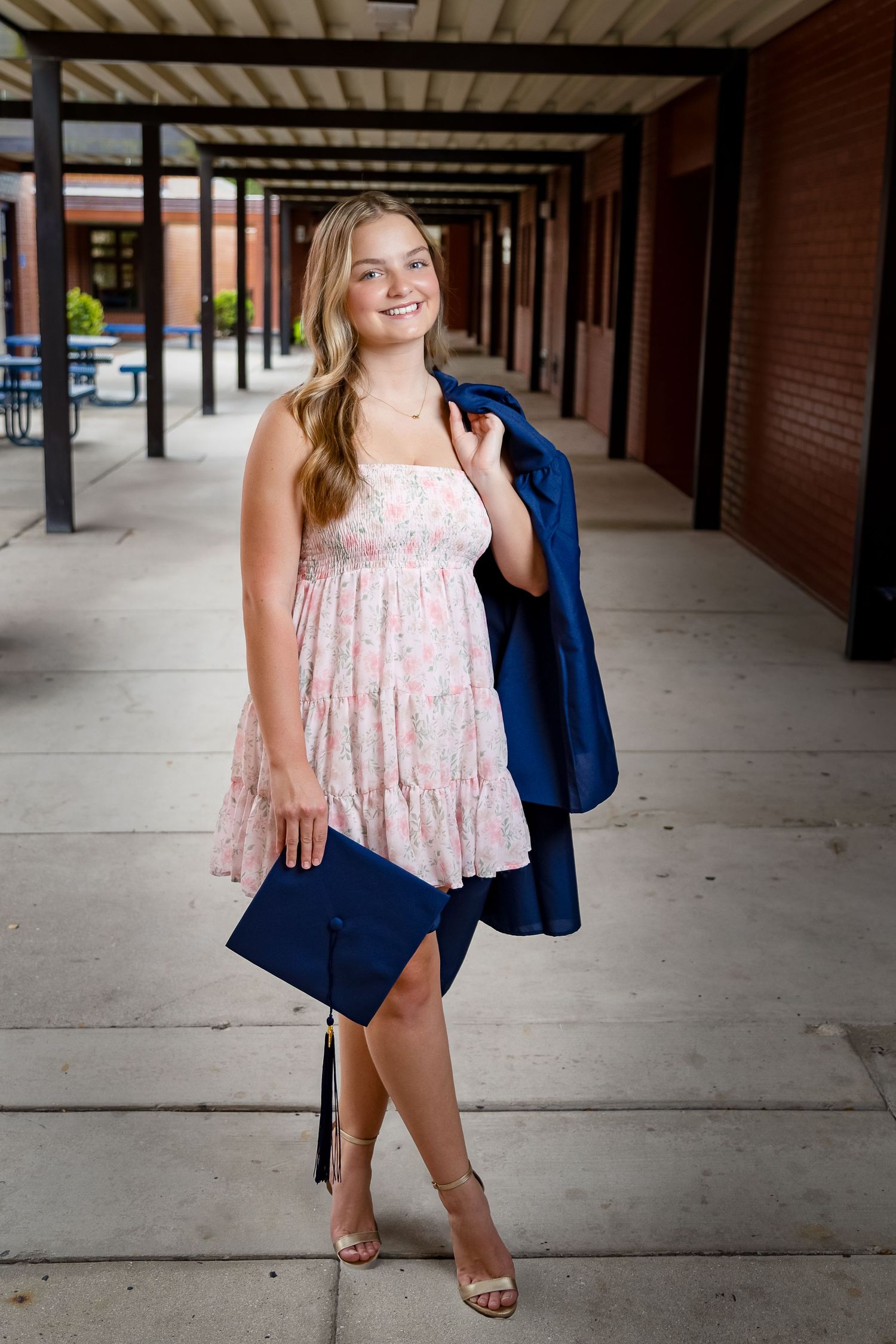 A person wearing a floral dress and navy blazer poses in a covered outdoor walkway with brick columns.